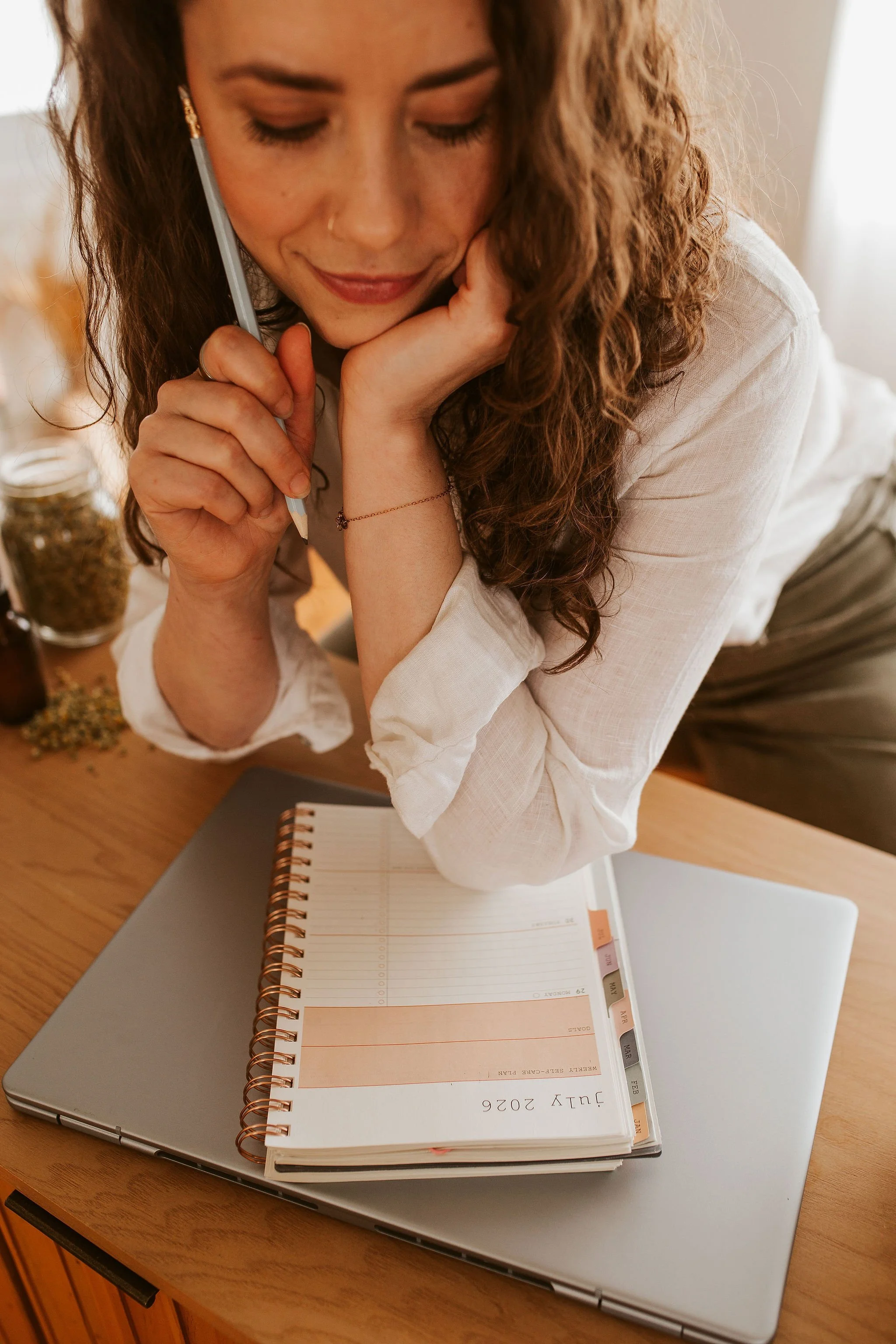 Emily LeDuc, Marketing Strategist, A woman with curly hair sitting at a wooden desk, looking down at her planner, with a laptop underneath the planner, and talking on the phone.