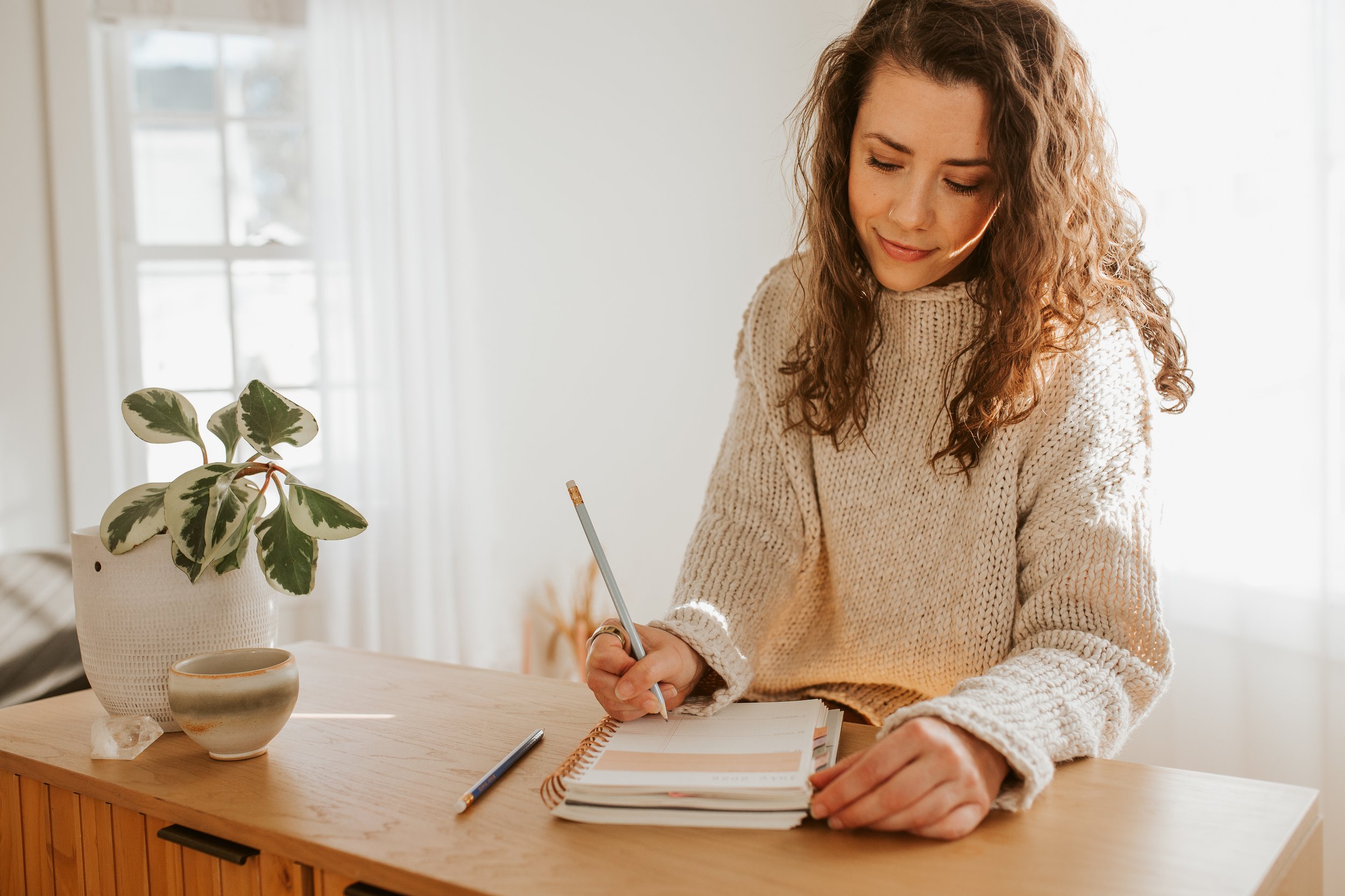 Emily LeDuc, Marketing Strategist, A woman with curly hair wearing a knit sweater, writing in a planner on a wooden table next to a potted plant and a mug.