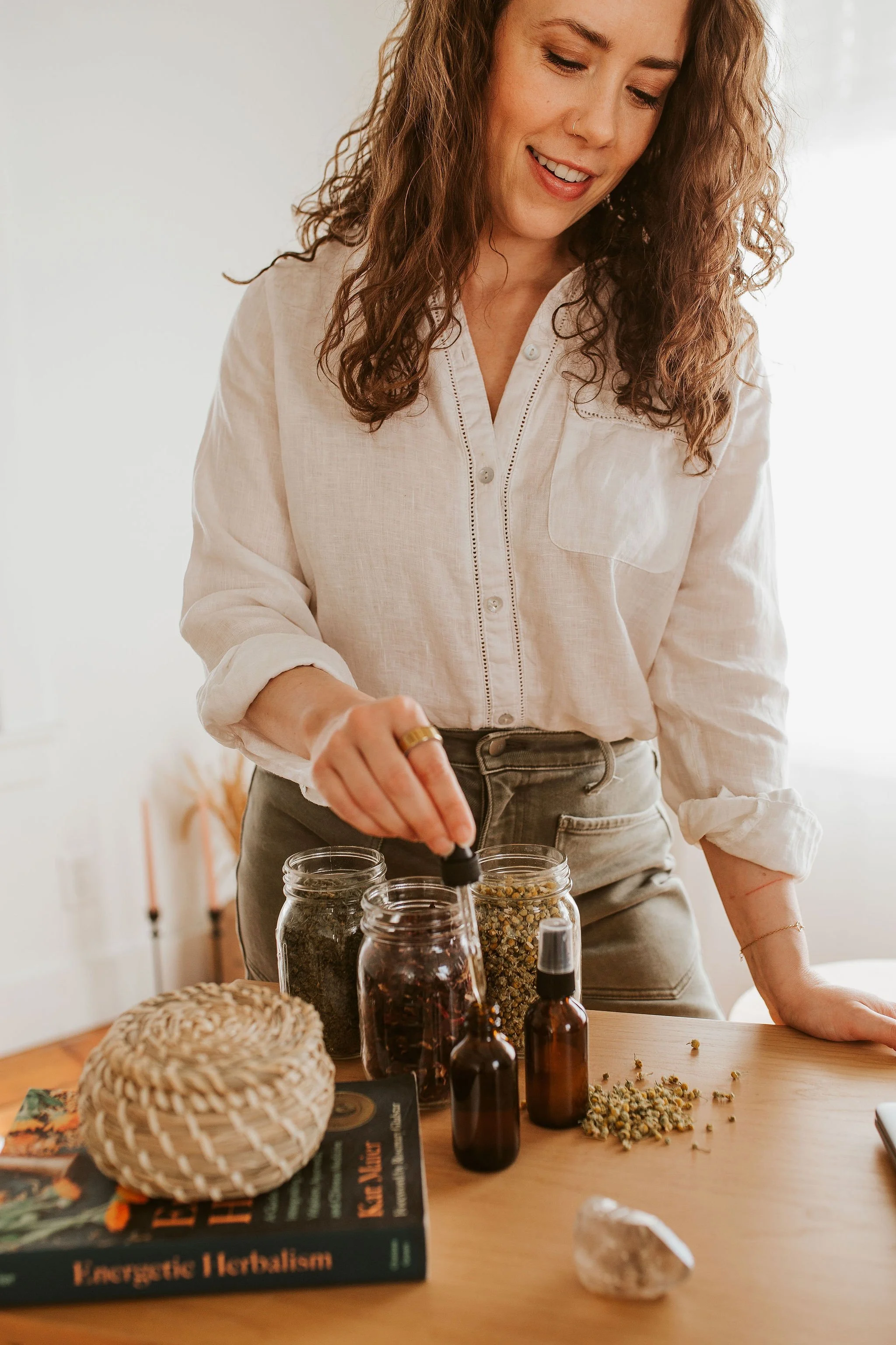 Emily LeDuc, Marketing Strategist, A woman with curly hair, wearing a cream-colored blouse, is preparing herbal remedies with dried herbs, essential oils, and books on herbalism on a wooden table.
