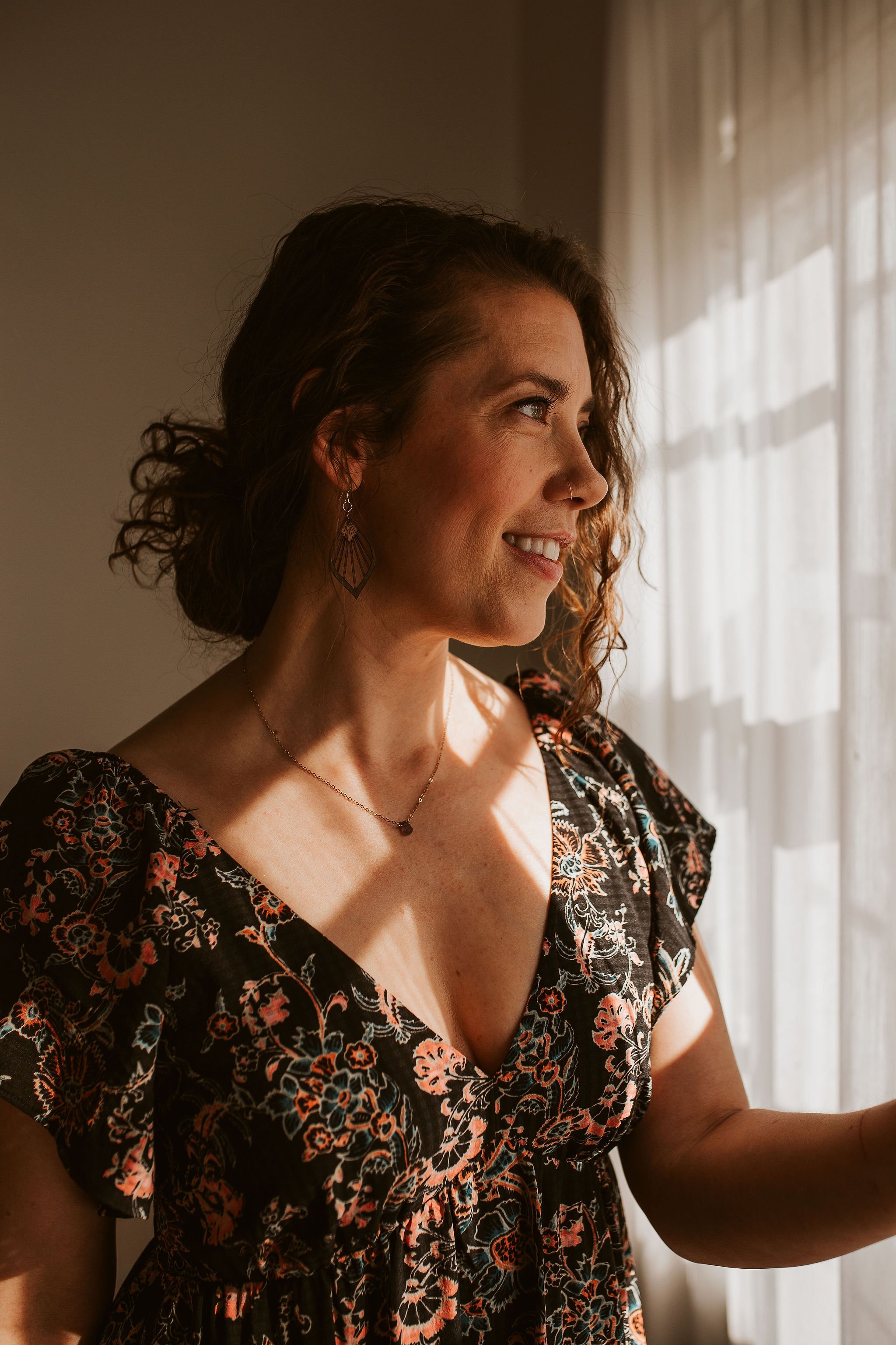 Emily LeDuc, A woman with curly hair smiling while looking out a window with sheer curtains.