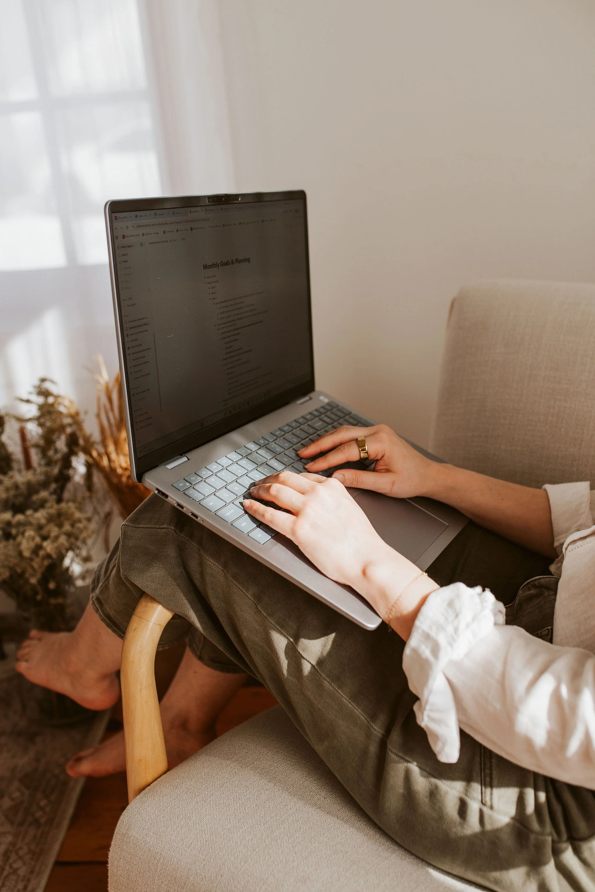Person sitting on a beige chair with legs crossed, using a silver laptop on their lap, in a bright room with light coming through a window. They are wearing a white shirt with rolled-up sleeves, dark jeans, a finger ring, and a bracelet.