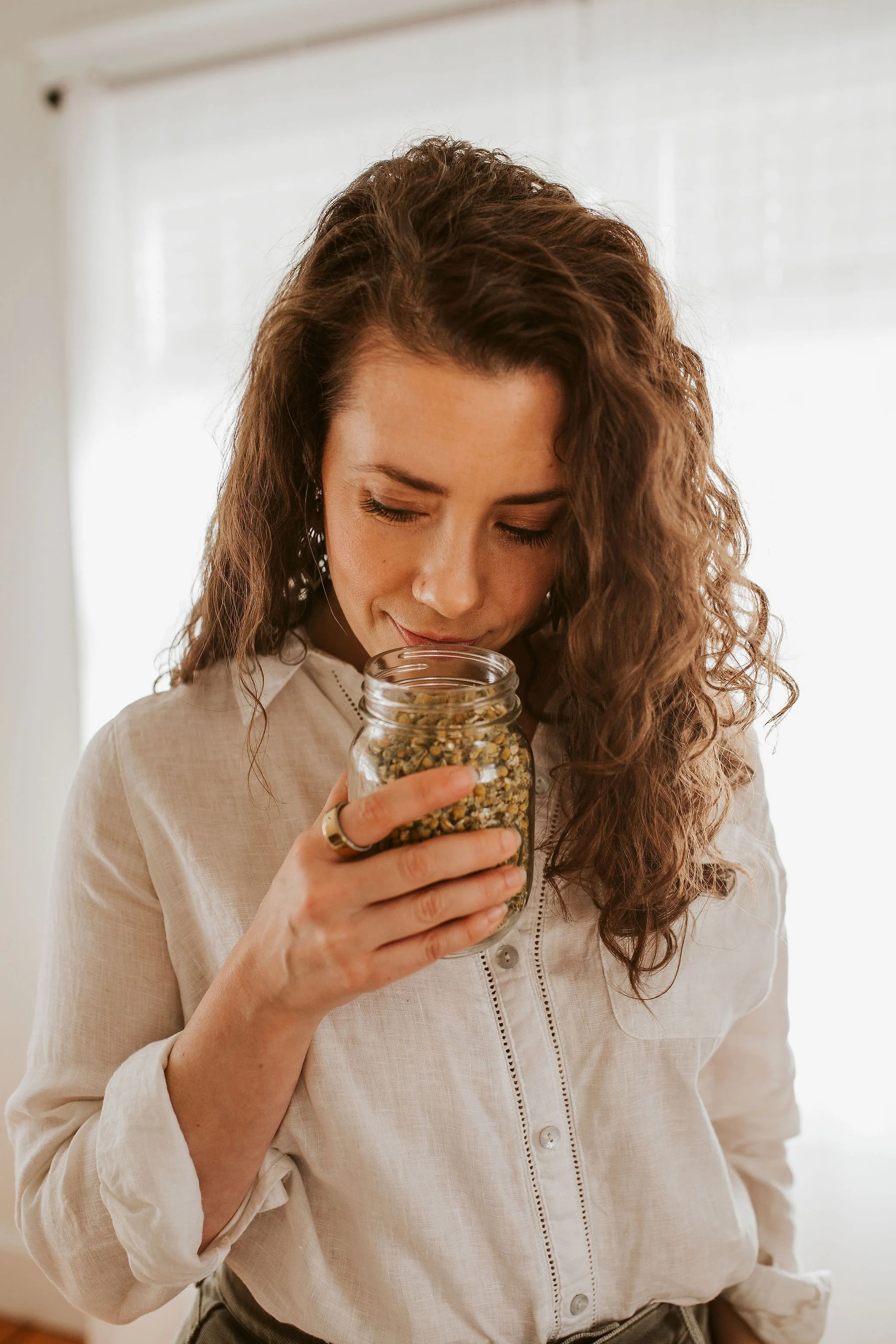 Emily LeDuc, Marketing Strategist, A woman with long, curly brown hair holding a glass jar of dried herbs or tea leaves close to her face, smelling it with her eyes closed.