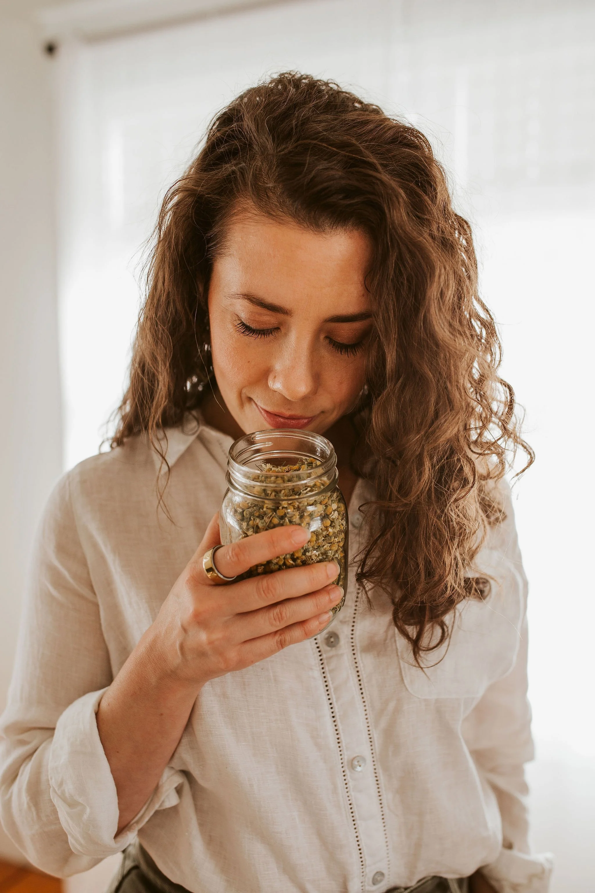 Emily LeDuc, Marketing Strategist, A woman with curly hair in a light-colored shirt holding a jar of herbal tea close to her face, smelling it.