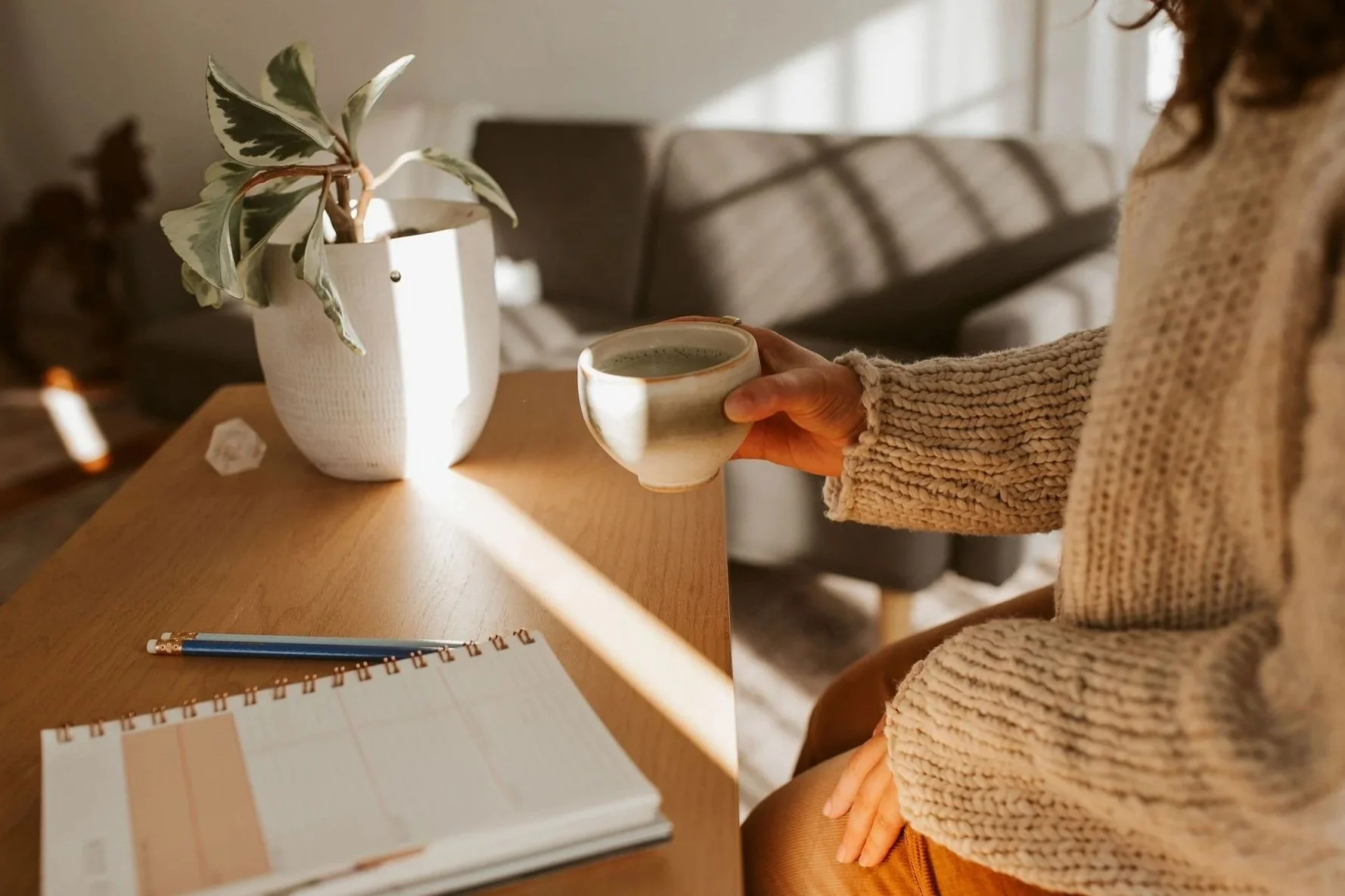 Person in a beige knitted sweater holding a ceramic cup of coffee or tea while sitting at a wooden table with a white potted plant, a notebook, and a pen. Sunlight streams into the room, casting shadows.
