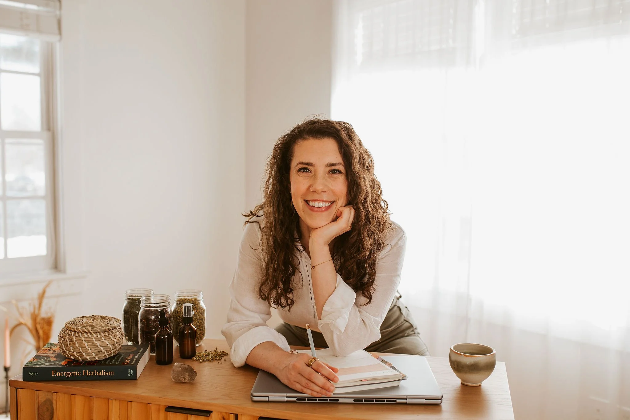 Emily LeDuc, Marketing Strategist, A woman with curly brown hair smiling while leaning on a desk with her chin resting on her hand, surrounded by herbal jars, a notebook, and a laptop in a bright room with daylight filtering through the curtains.