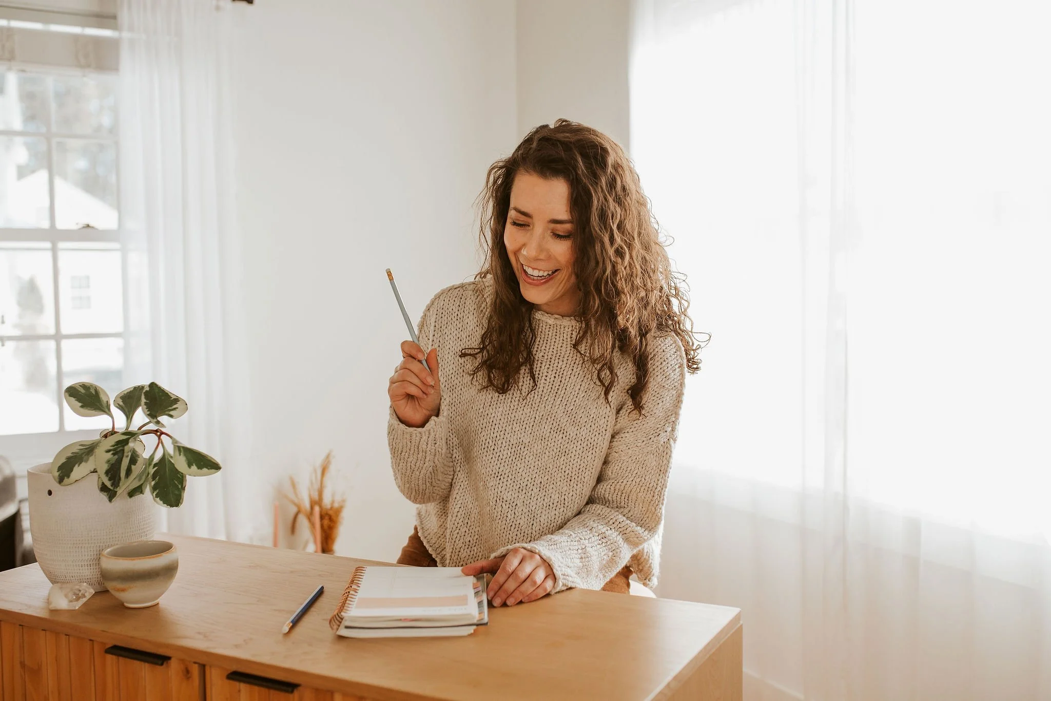 Emily LeDuc, Marketing Strategist, A woman with curly hair in a beige sweater smiling and holding a pencil in a bright room with sheer curtains, a potted plant, and an open notebook on a wooden desk.