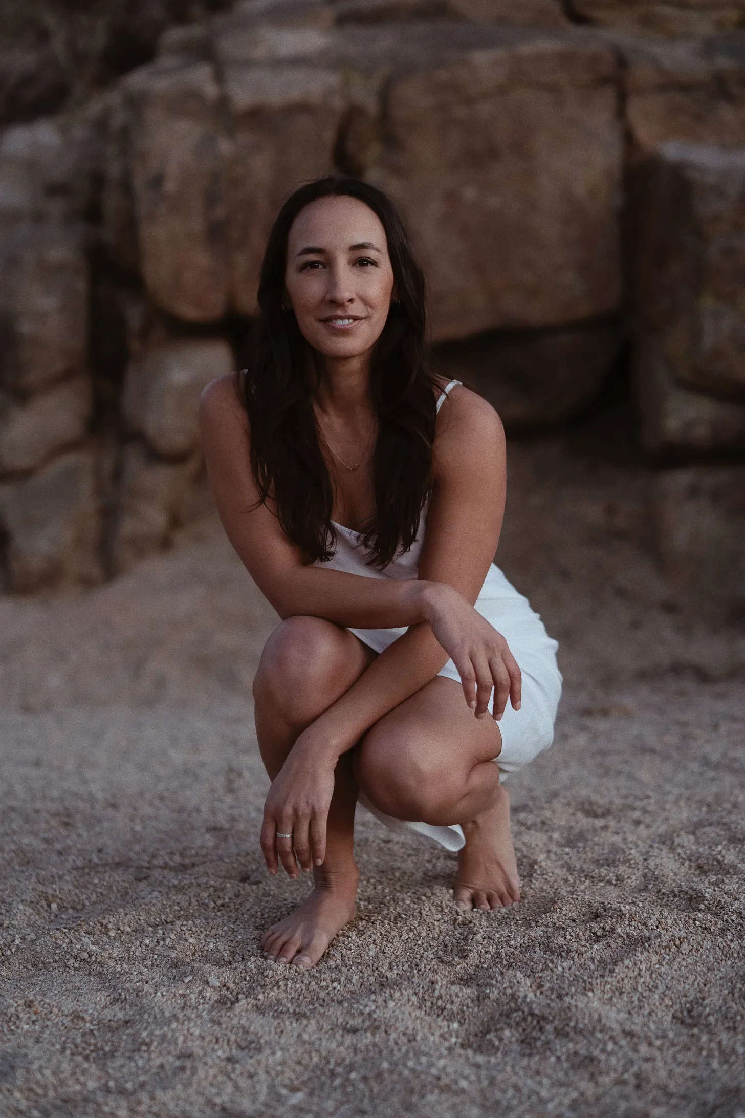 A woman with long dark hair, wearing a white sleeveless dress, is squatting on sandy ground with a stone wall in the background, smiling gently.