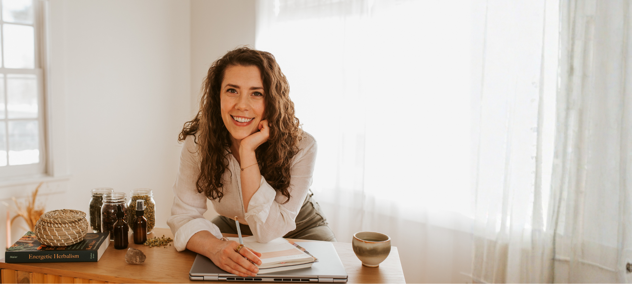 A woman with curly brown hair smiling while sitting at a desk, holding a pen over an open notebook. The desk has various jars, herbal bottles, a book titled 'Energetic Herbalism,' a small woven basket, a rock, and a ceramic mug. The room has natural light coming through a window with sheer curtains.