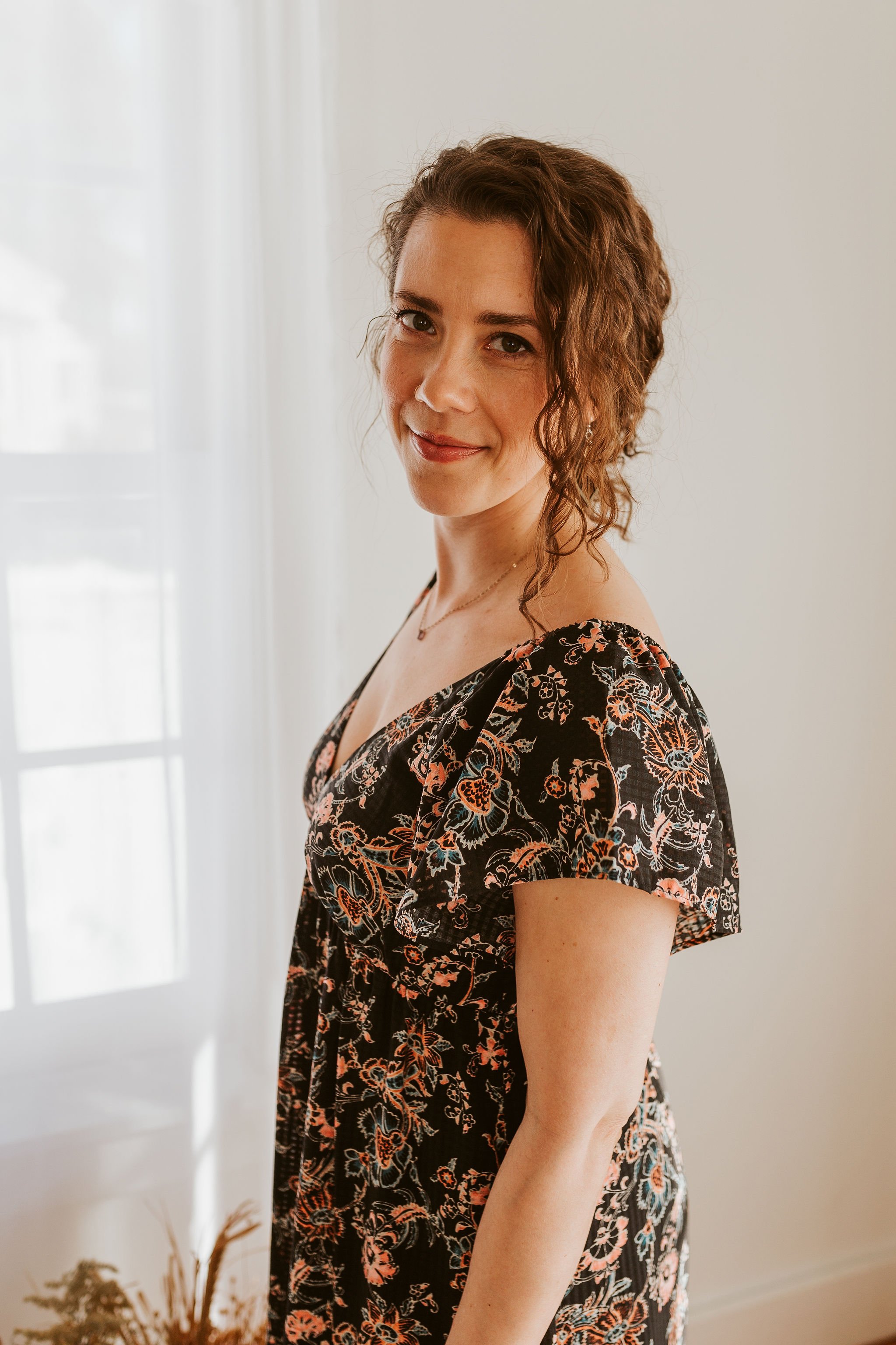 Emily LeDuc, Marketing Strategist, A woman with curly brown hair, wearing a black floral dress, standing in front of a window with white curtains in a bright room.