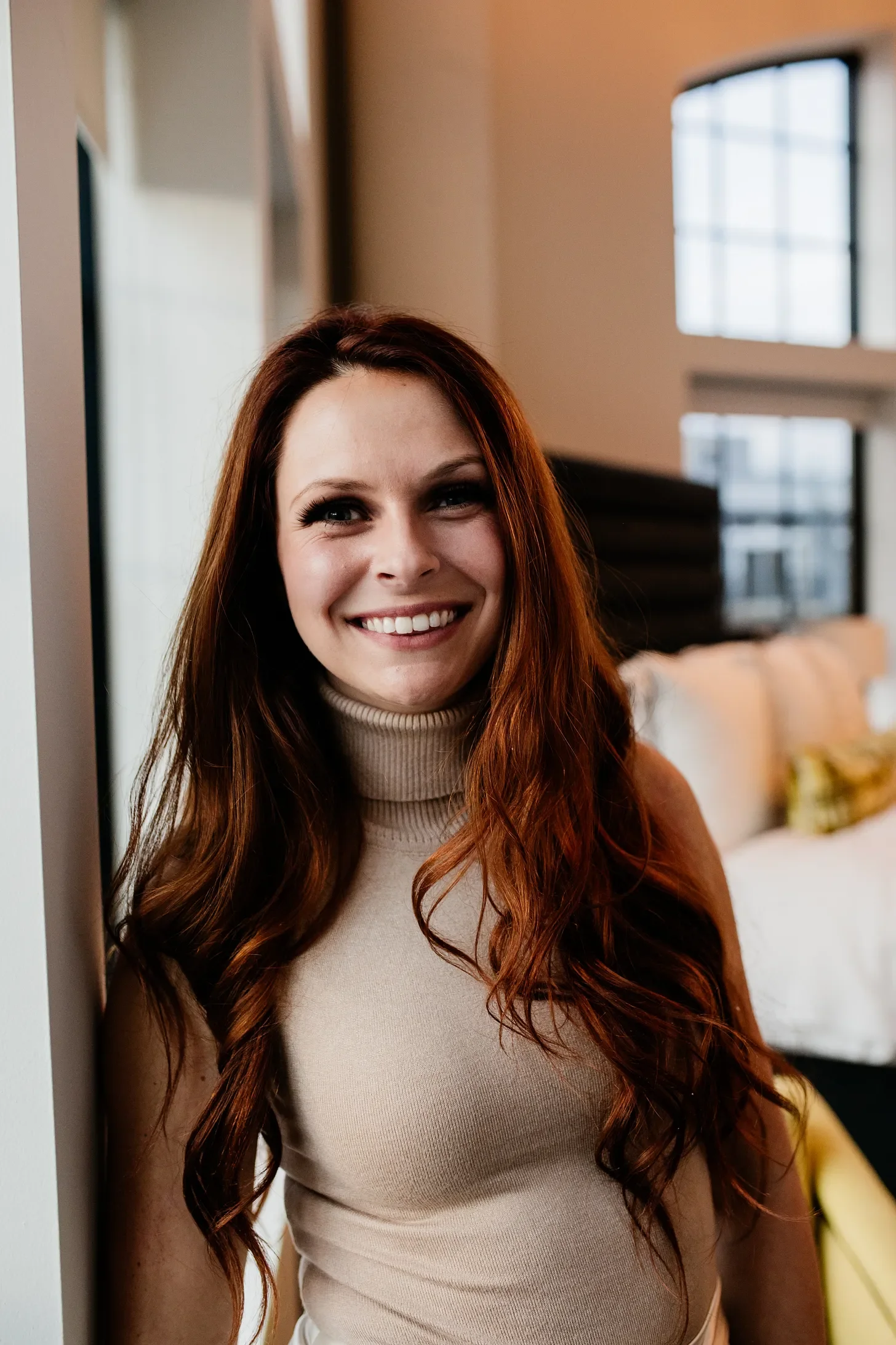 A smiling woman with long red hair wearing a beige turtleneck, standing indoors near a wall, with large windows and a sofa in the background.