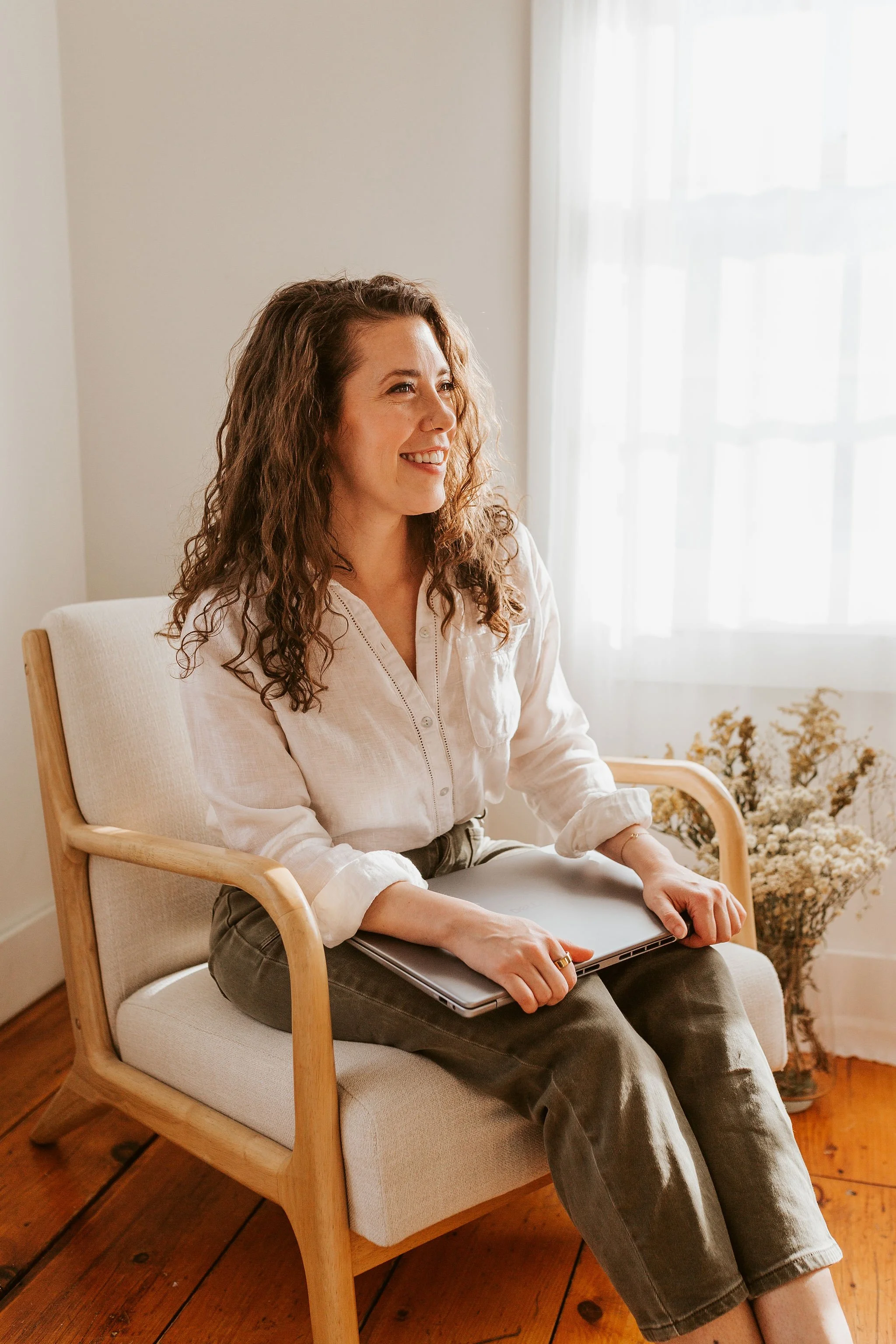 Emily LeDuc, Marketing Strategist, A woman sitting on a light-colored cushioned armchair by a window with sheer curtains, holding a laptop on her lap, smiling, with a vase of dried flowers on the floor beside her.