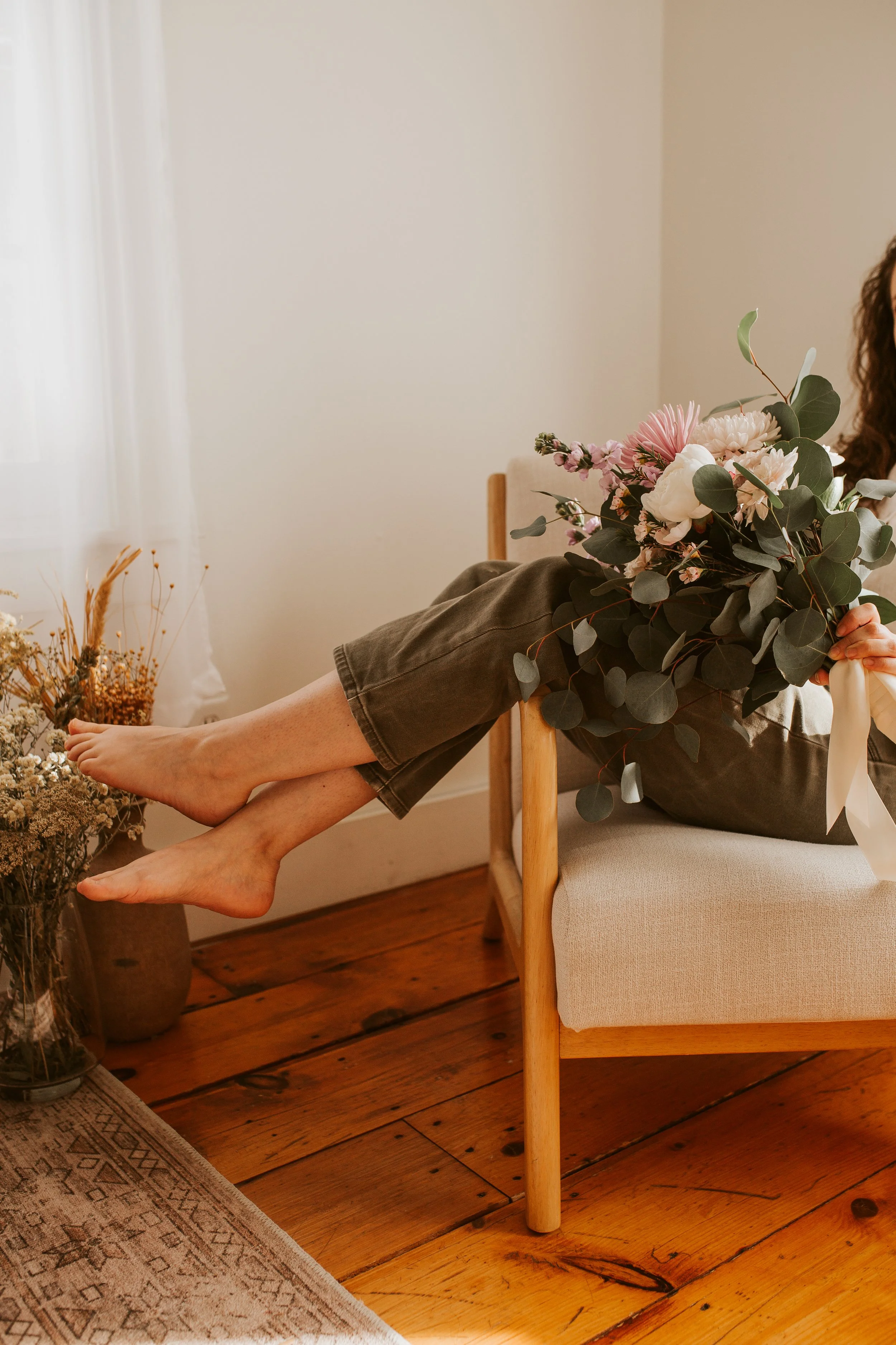 Person sitting on a wooden chair, holding a bouquet of flowers, with bare feet resting on an adjoining flower arrangement, in a cozy room.