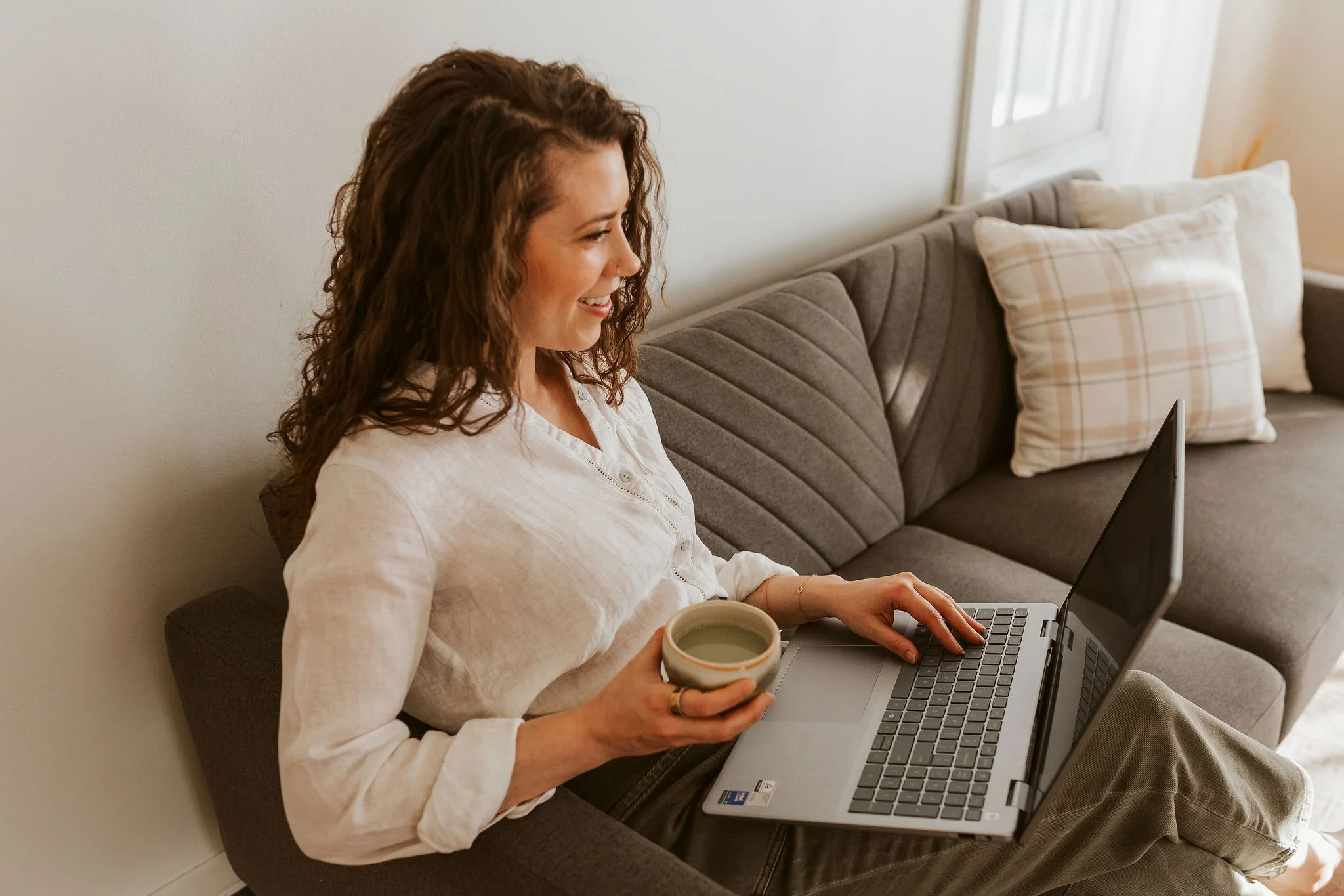 Emily LeDuc, Marketing Strategist, A woman with curly brown hair sitting on a gray sofa, working on a laptop, holding a mug, smiling, in a well-lit room.