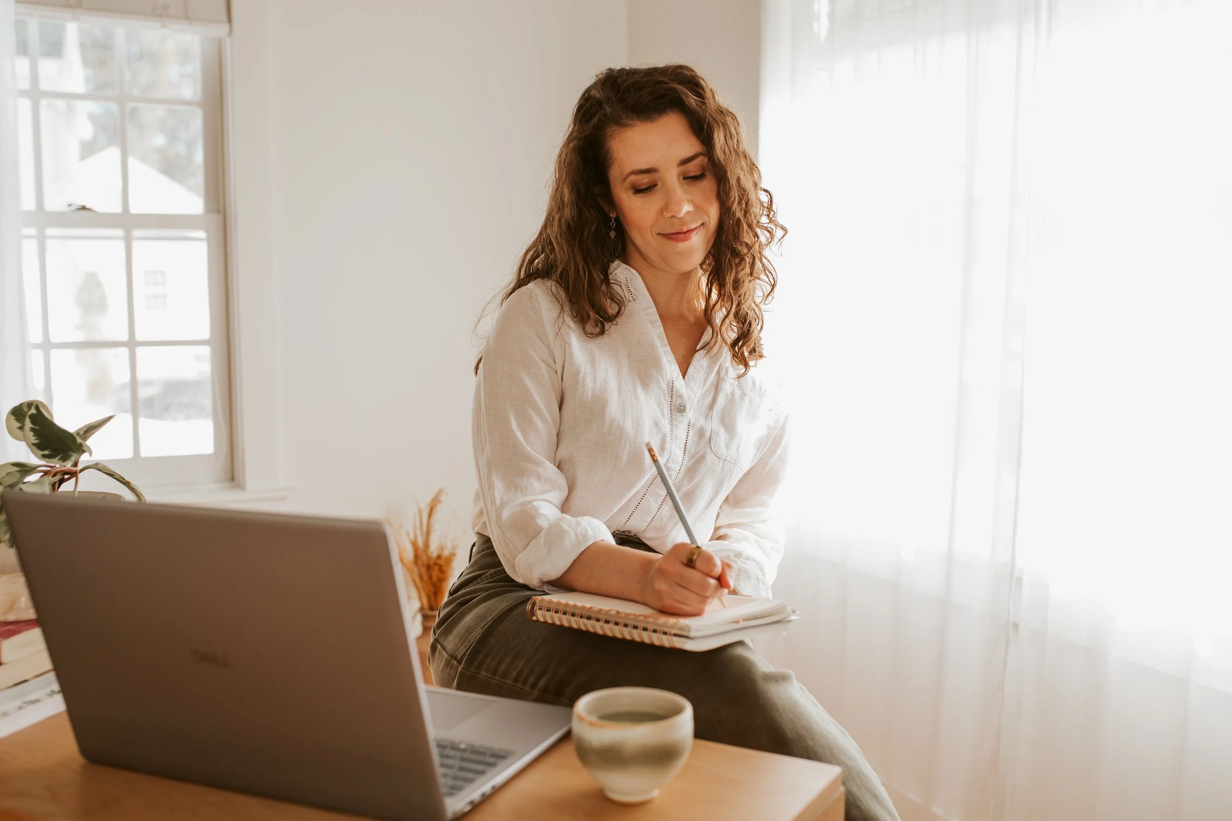 Emily LeDuc, Marketing Strategist, A woman with curly brown hair wearing a white blouse, sitting at a wooden table with a laptop, notebook, pen, and a cup of coffee in a bright room with white curtains.