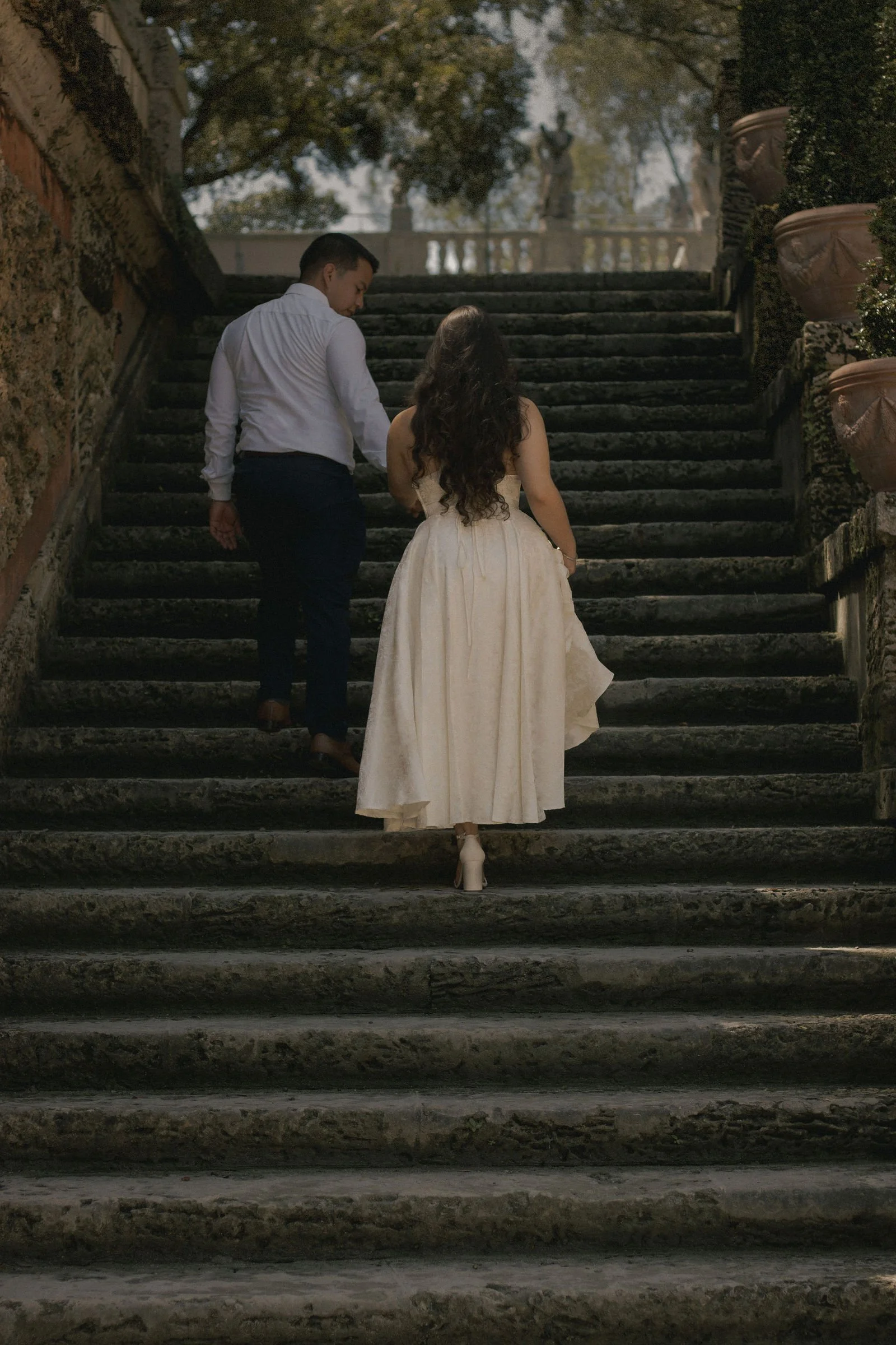 A man and woman walking up outdoor stone steps, with the woman in a white dress and the man in a white shirt and dark pants, surrounded by large flower pots and greenery.