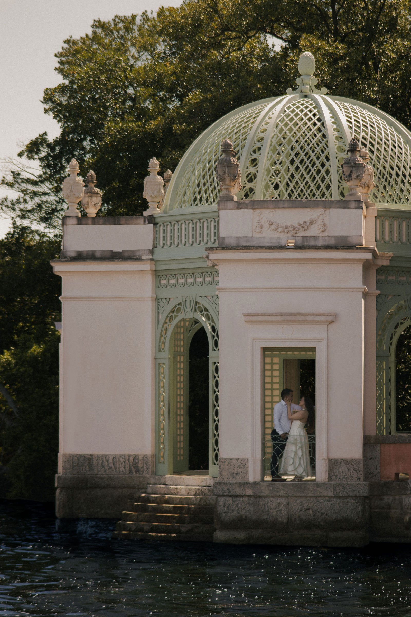 A couple standing inside a white pavilion with a green roof and decorative lanterns, overlooking a body of water, with trees in the background.