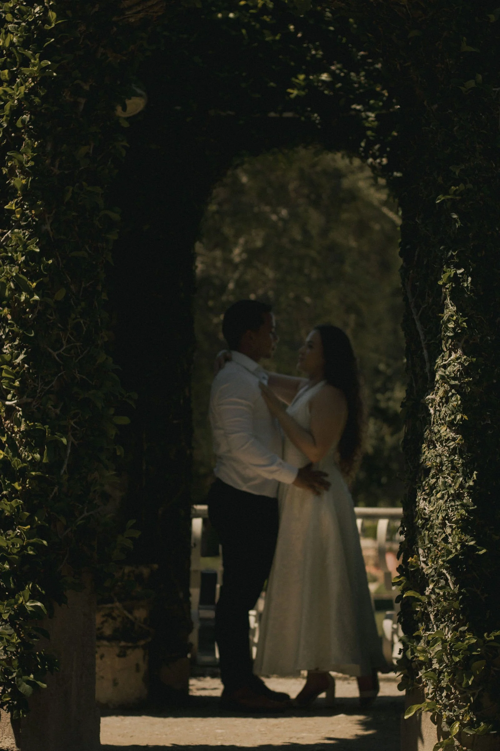 A couple dancing closely beneath a tree archway in a park during sunset, with the man wearing a white shirt and dark pants, and the woman in a white dress.