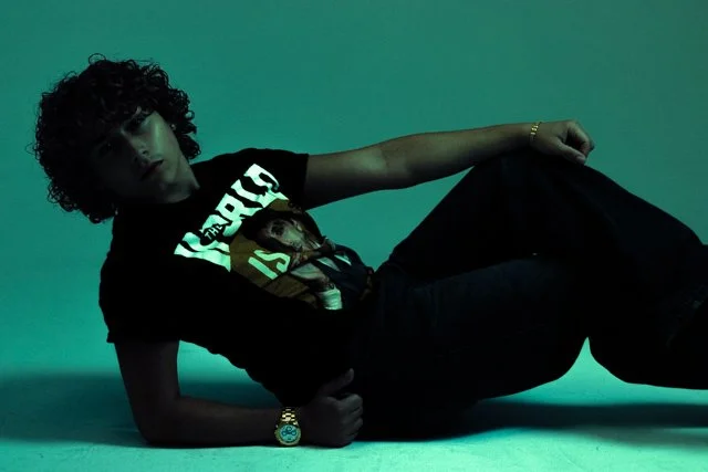 A man with curly hair lying on the floor against a green background, wearing a black graphic T-shirt, black pants, and a wristwatch.