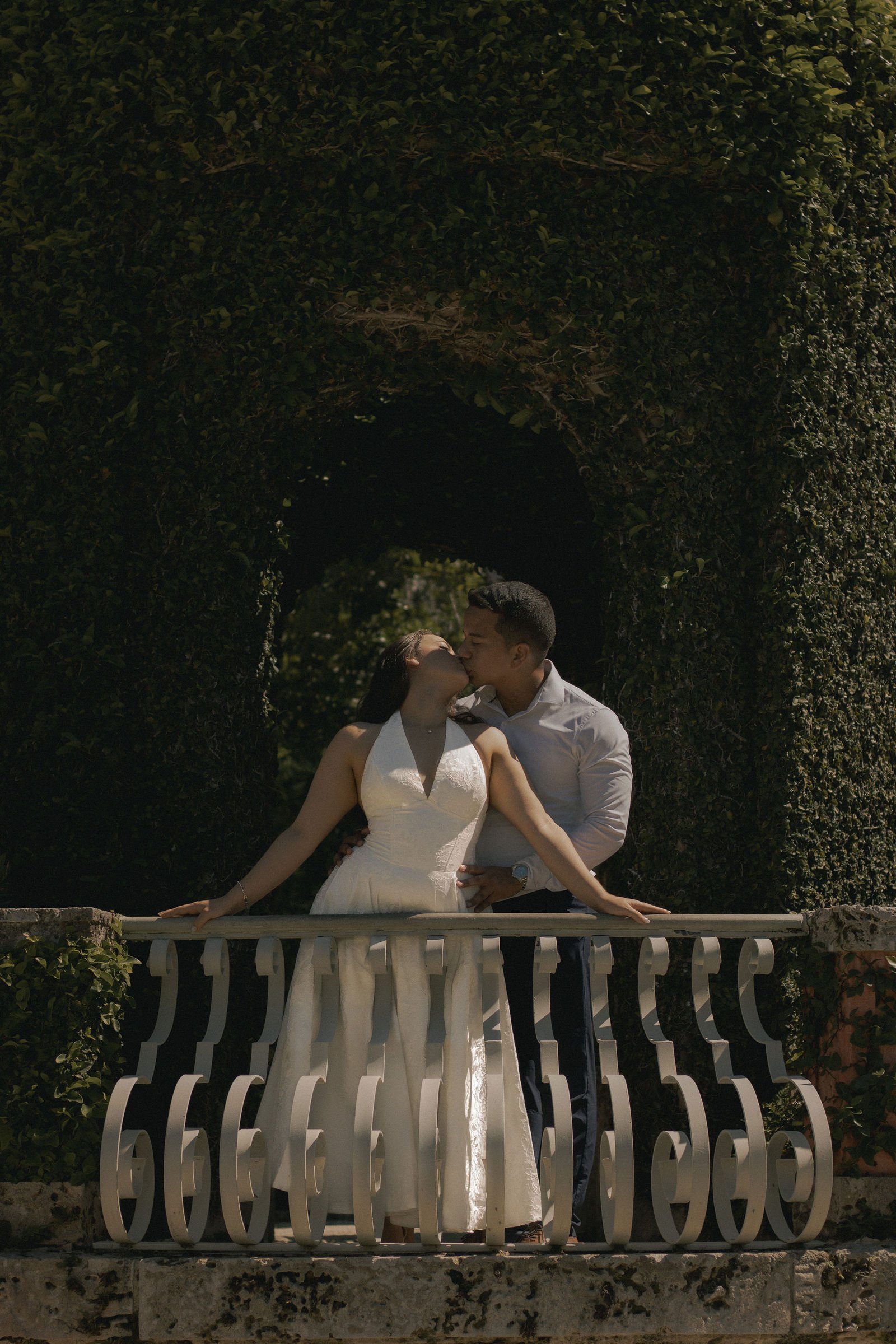 A couple kissing while standing on a balcony with a white railing, surrounded by greenery and trees.
