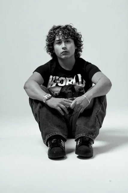 Young man with curly hair sitting on the floor with his arms resting on his knees, wearing a black T-shirt with a graphic, jeans, and sneakers, against a plain light background.