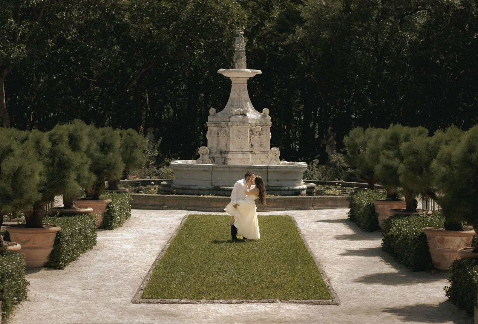 A bride and groom sharing a kiss in a garden with a large white marble fountain behind them, surrounded by potted plants and trees.