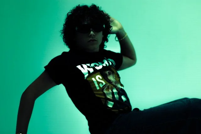 Person with curly hair wearing sunglasses, a black graphic T-shirt, sitting on a surface against a green-lit background.