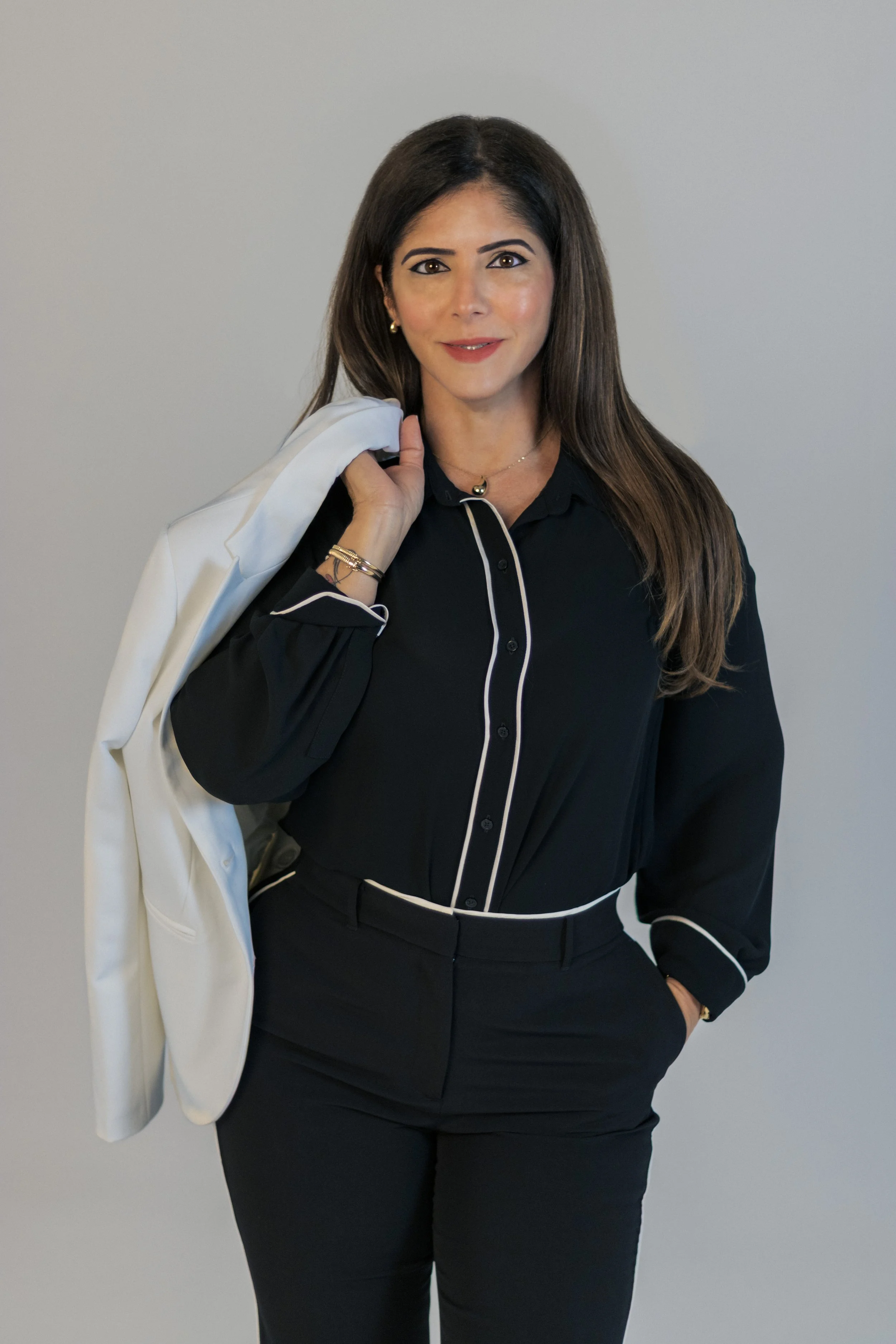 Woman with long brown hair in black outfit with white piping, holding white blazer over her shoulder, standing against a gray background.