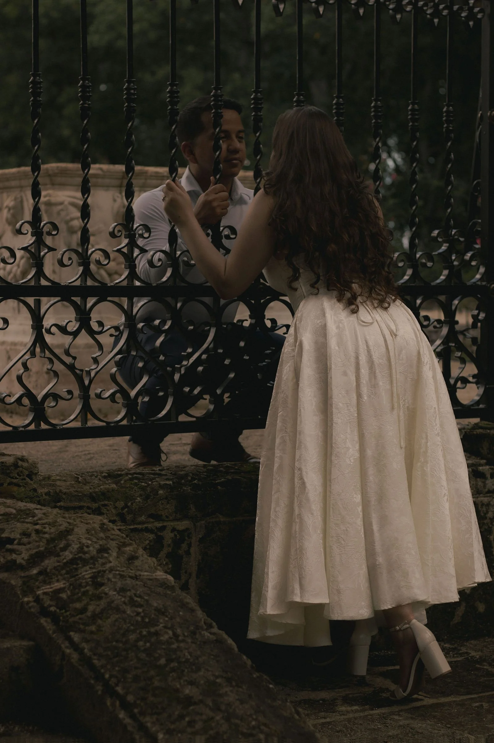 A young woman in a white vintage-style dress with long curly hair stands behind a black wrought iron gate, gazing at a young man sitting on the other side, in an outdoor park setting during dusk.