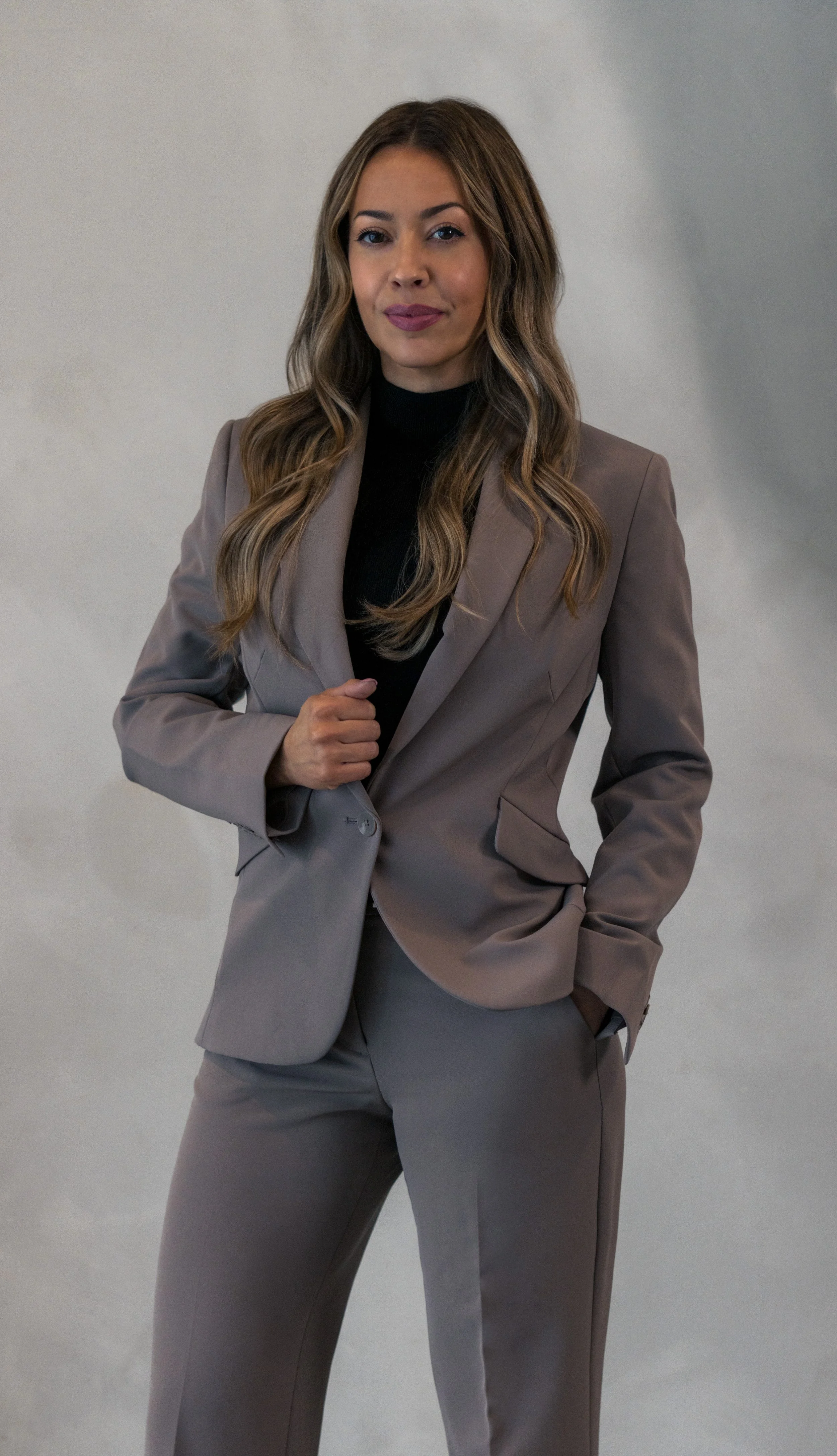 A confident woman with long, wavy hair in a taupe pantsuit and black top, standing against a plain light gray background.
