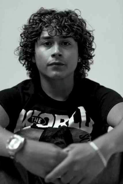 Black and white photo of a young man with curly hair wearing a graphic t-shirt, sitting with arms crossed, looking directly at the camera.