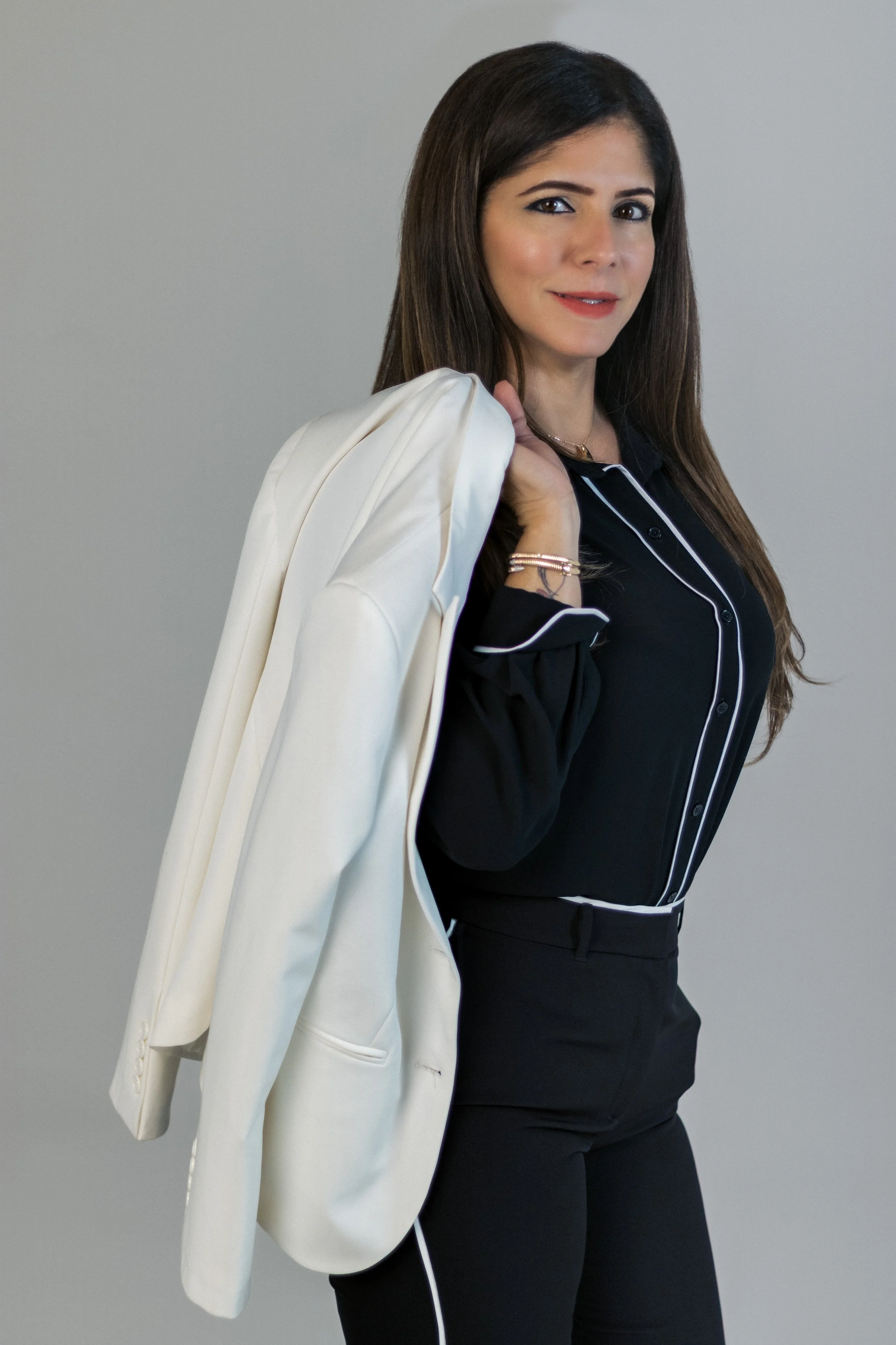A woman with long, dark hair is wearing a black outfit with white piping and holding a white blazer over her shoulder, standing against a plain gray background.