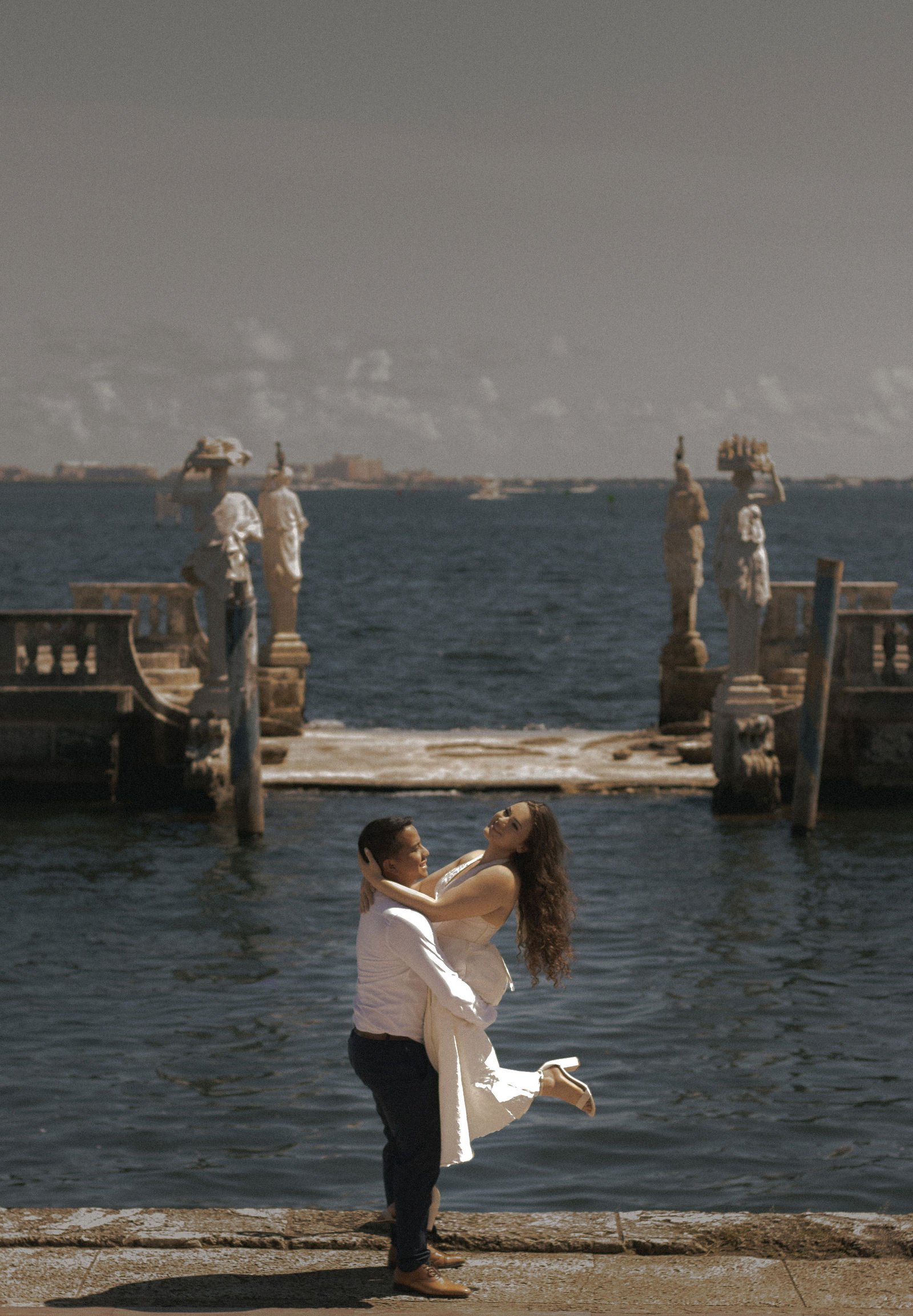 A couple dancing by the water, with the man lifting the woman in his arms, both smiling, with a pier and statues in the background.