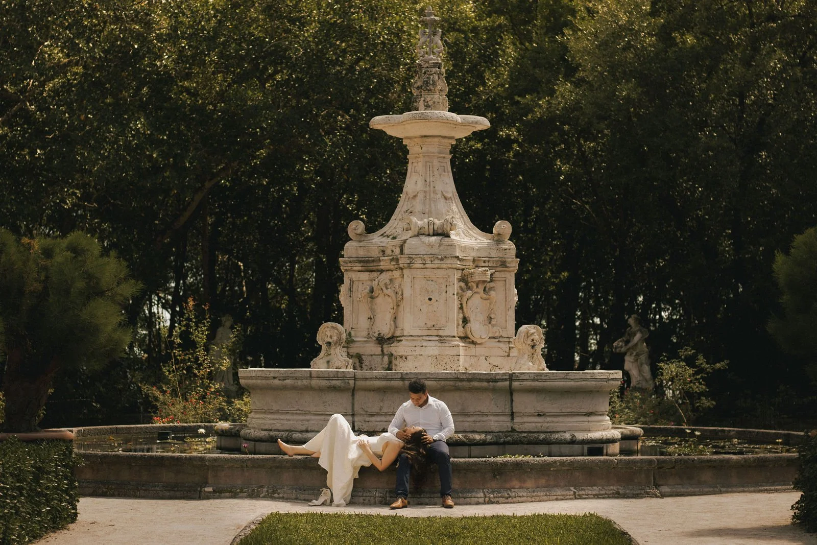 A couple sitting by a decorative stone fountain in a park, with the woman lying down with her head on the man's lap, surrounded by lush green trees.