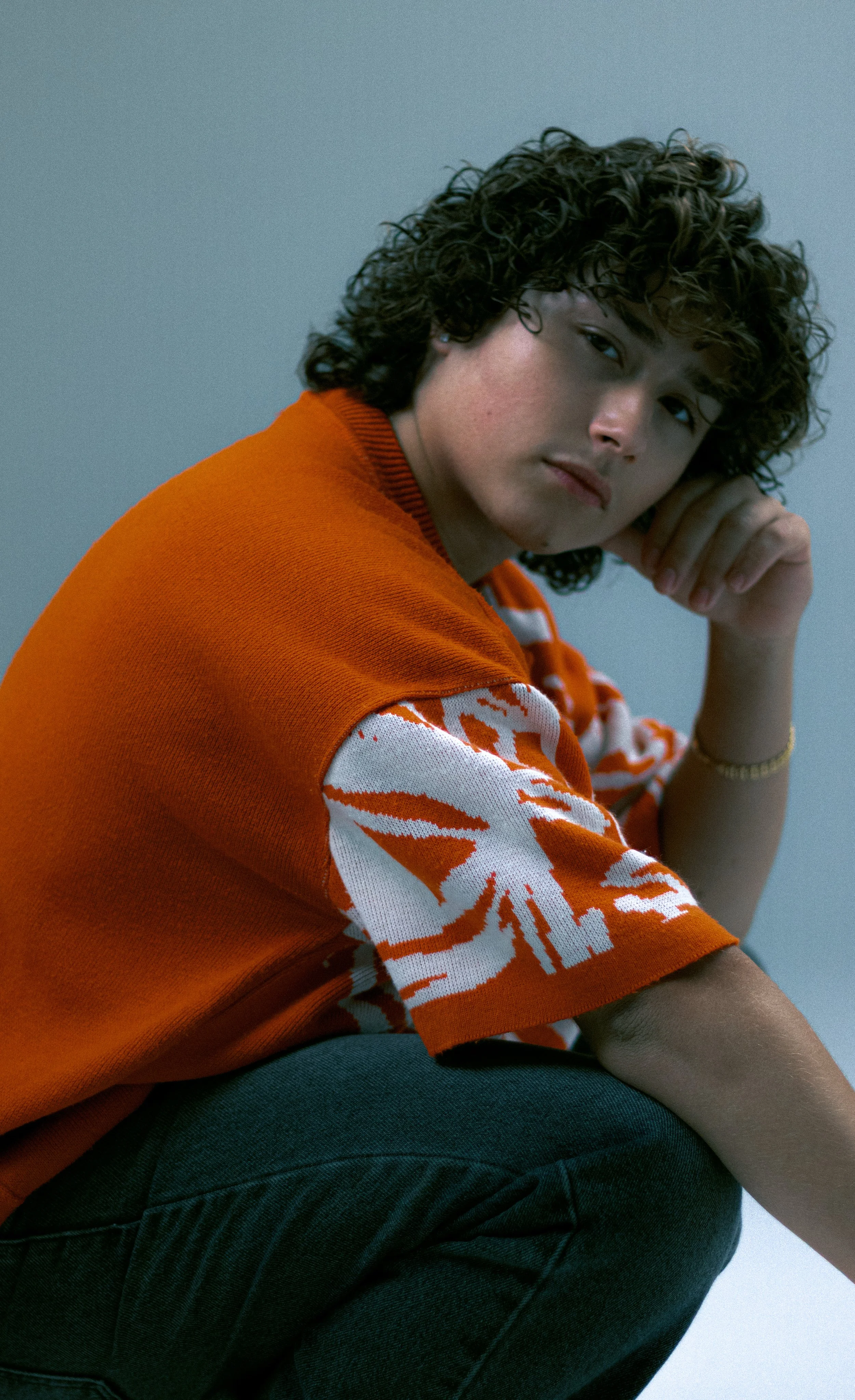 A young woman with curly dark hair wearing an orange and white patterned shirt and dark jeans, sitting and resting her head on her hand against a plain gray background.