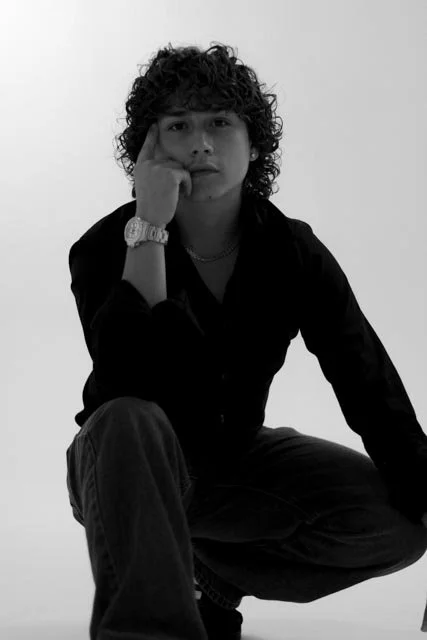 Black and white photo of a young man with curly hair, sitting with one leg up, resting his chin on his hand, wearing a dark shirt and a watch.
