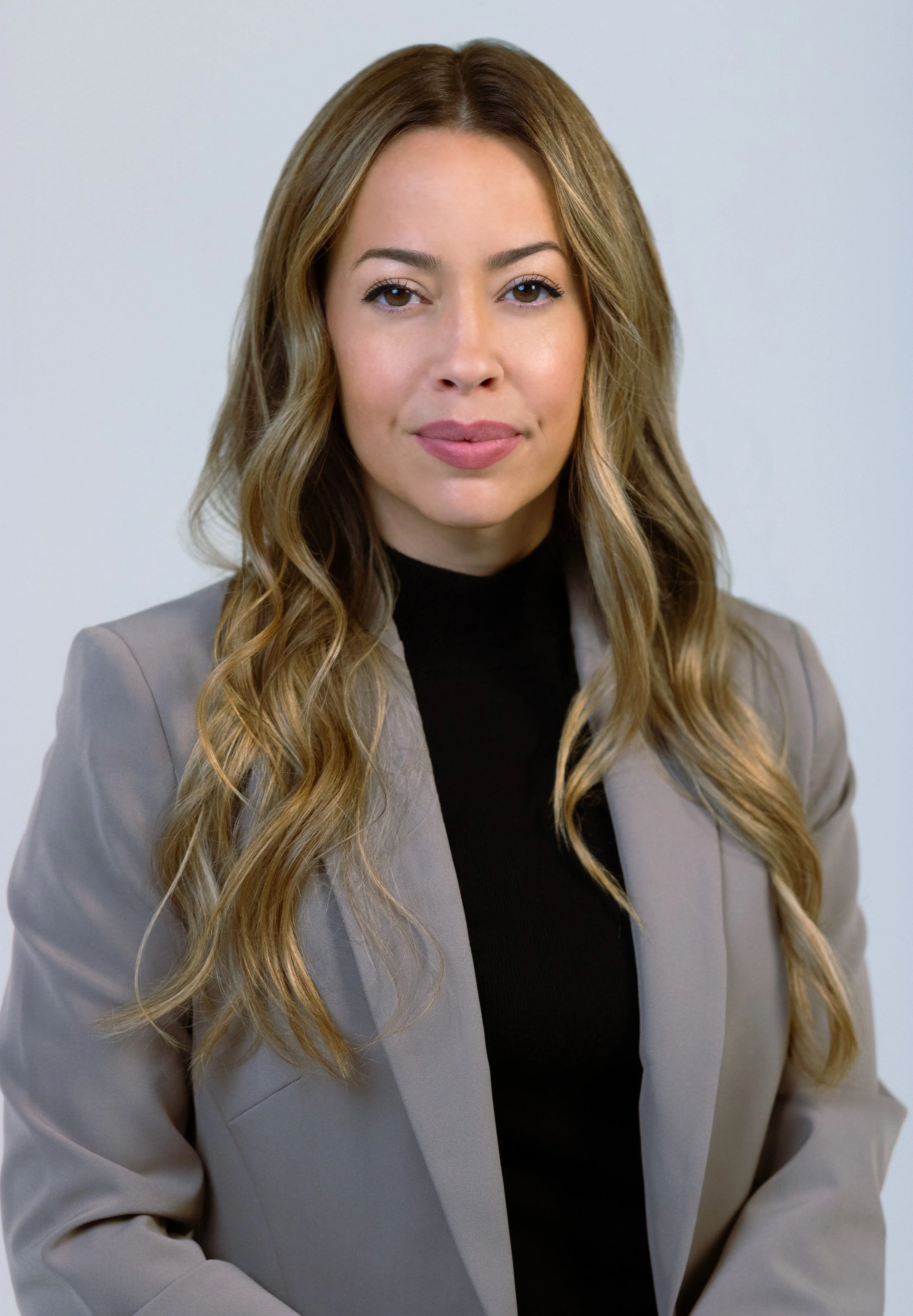 A woman with long, wavy blonde hair wearing a beige blazer over a black top.