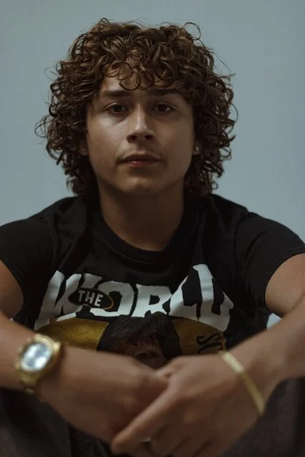A young man with curly brown hair and light skin sitting against a plain gray background.