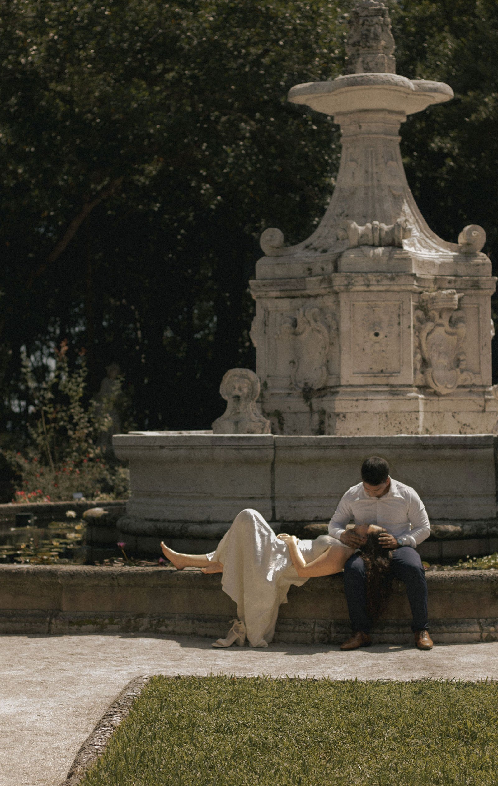 A couple is sitting on the edge of a fountain in a park at night. The woman, wearing a white dress, is lying back with her feet off the ground while the man, dressed in a white shirt and dark pants, is sitting upright, gently holding the woman's head