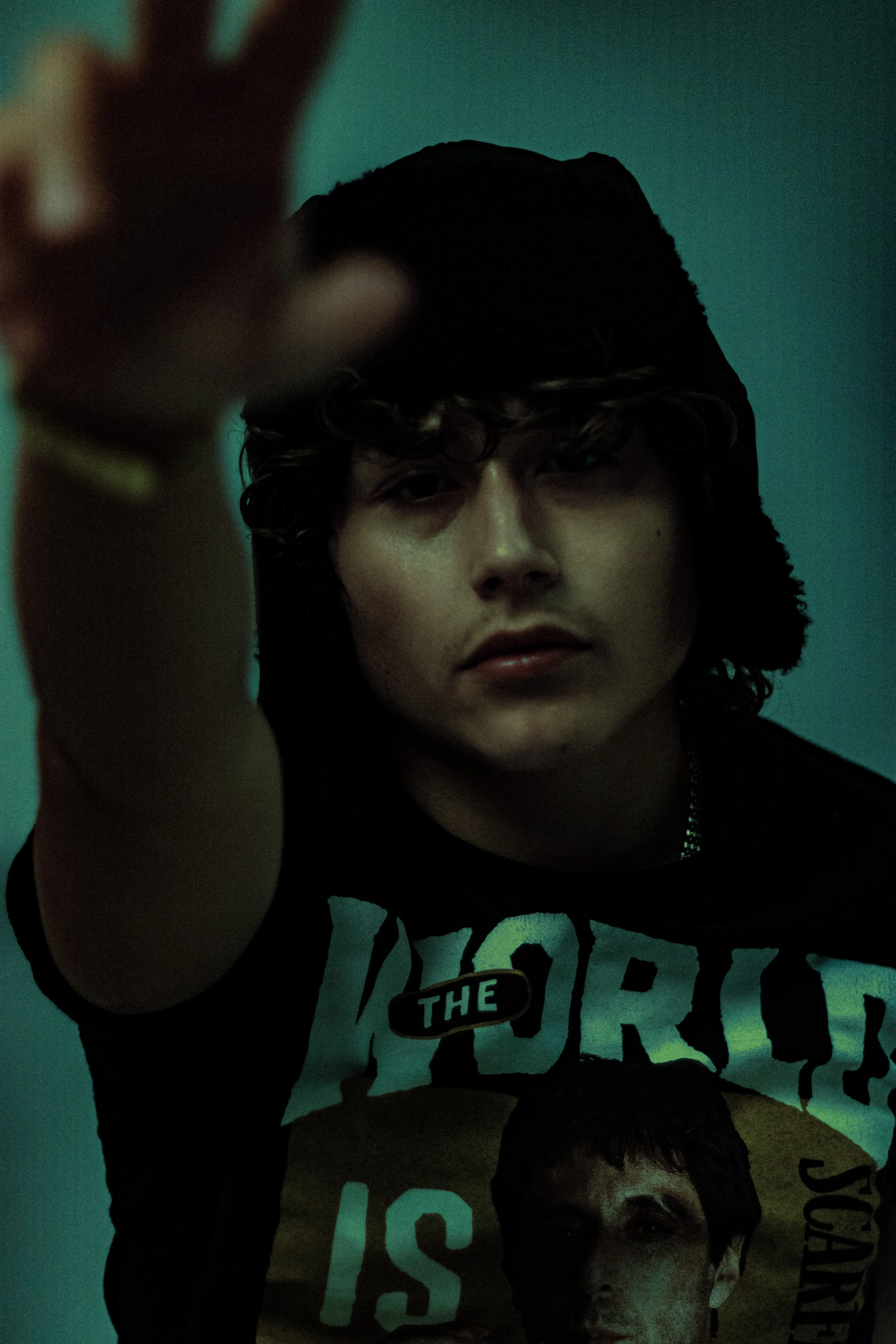 A person with curly hair and light skin poses with a peace sign, wearing a black hoodie, against a plain, teal-colored background.