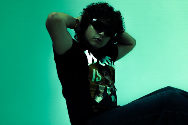 Person with curly hair wearing sunglasses and a black T-shirt sitting against a green background.