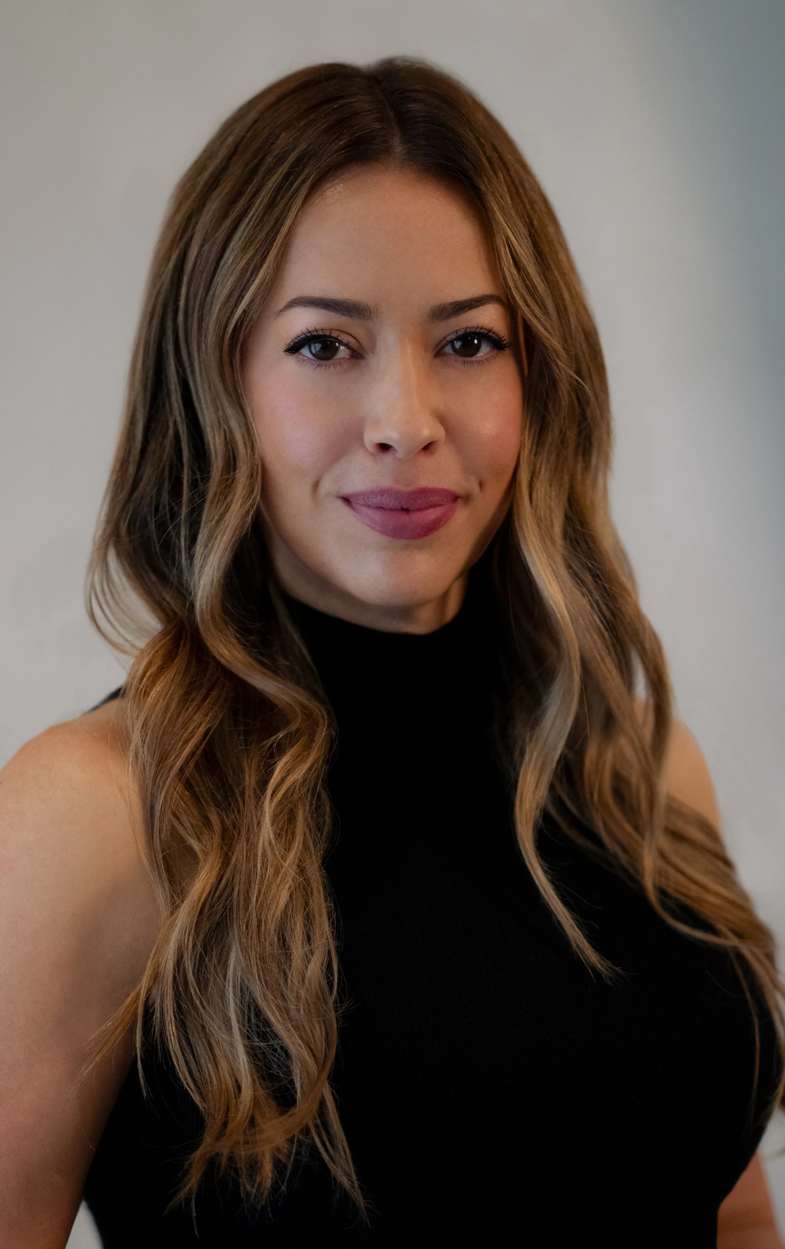 A woman with long wavy hair, wearing a black sleeveless top, smiling softly, against a neutral background.
