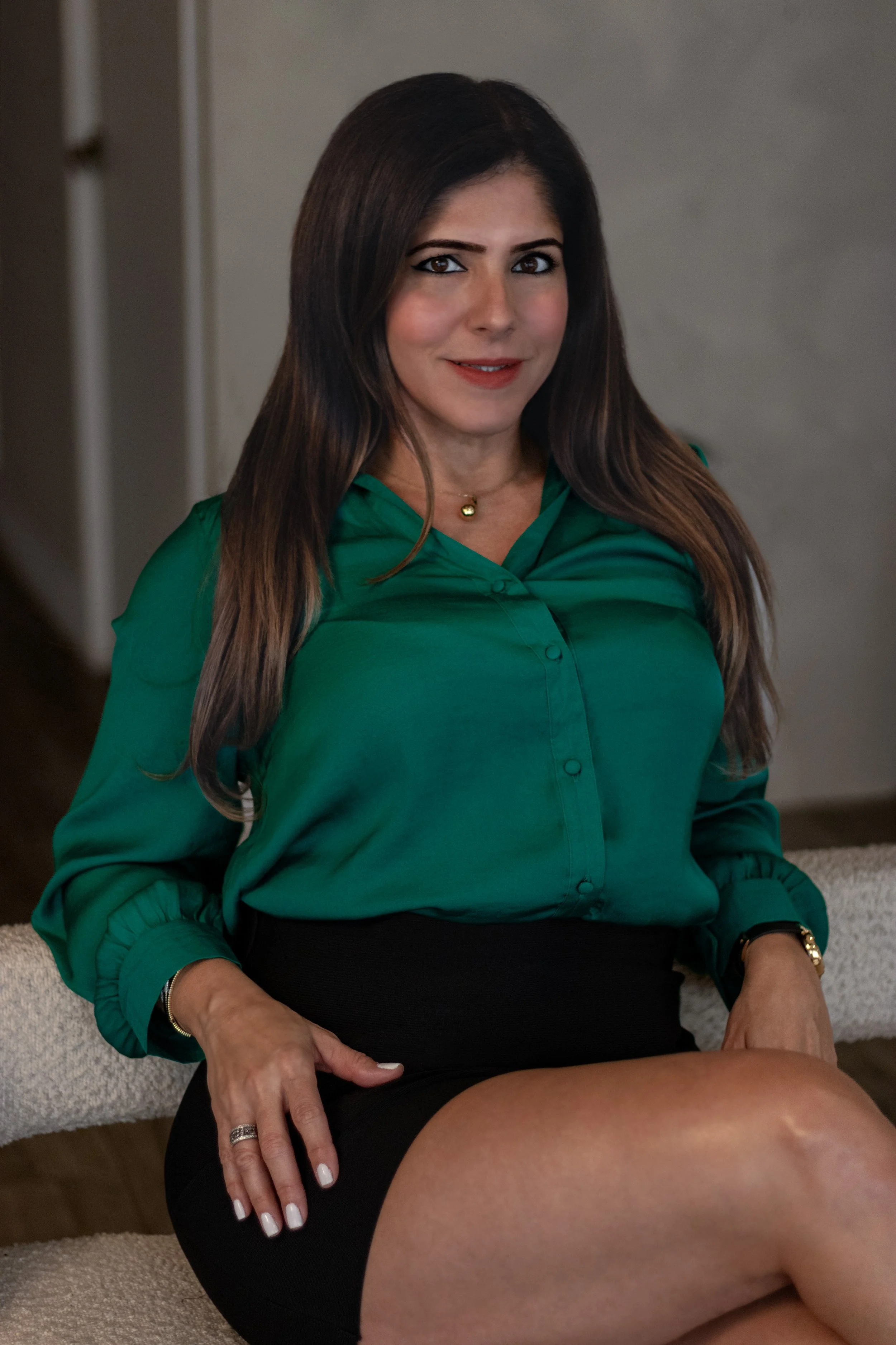 A woman with long brown hair wearing a green blouse and black skirt, sitting on a beige carpeted floor, in a room with neutral walls.