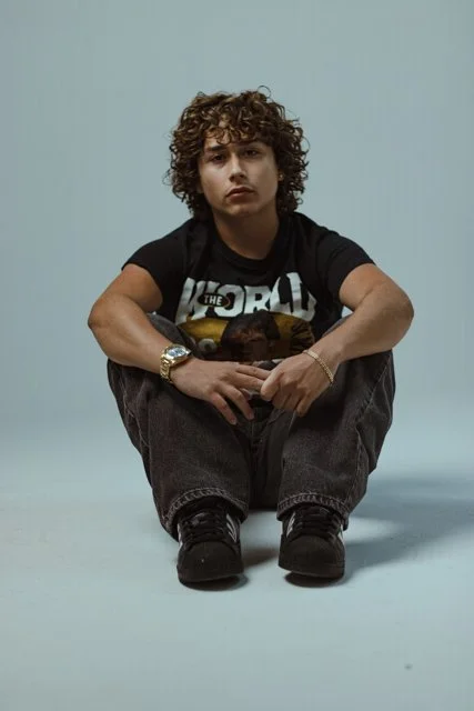 Young person with curly hair sitting on the floor against a plain light background, wearing a black t-shirt with graphic text, dark pants, and black shoes.