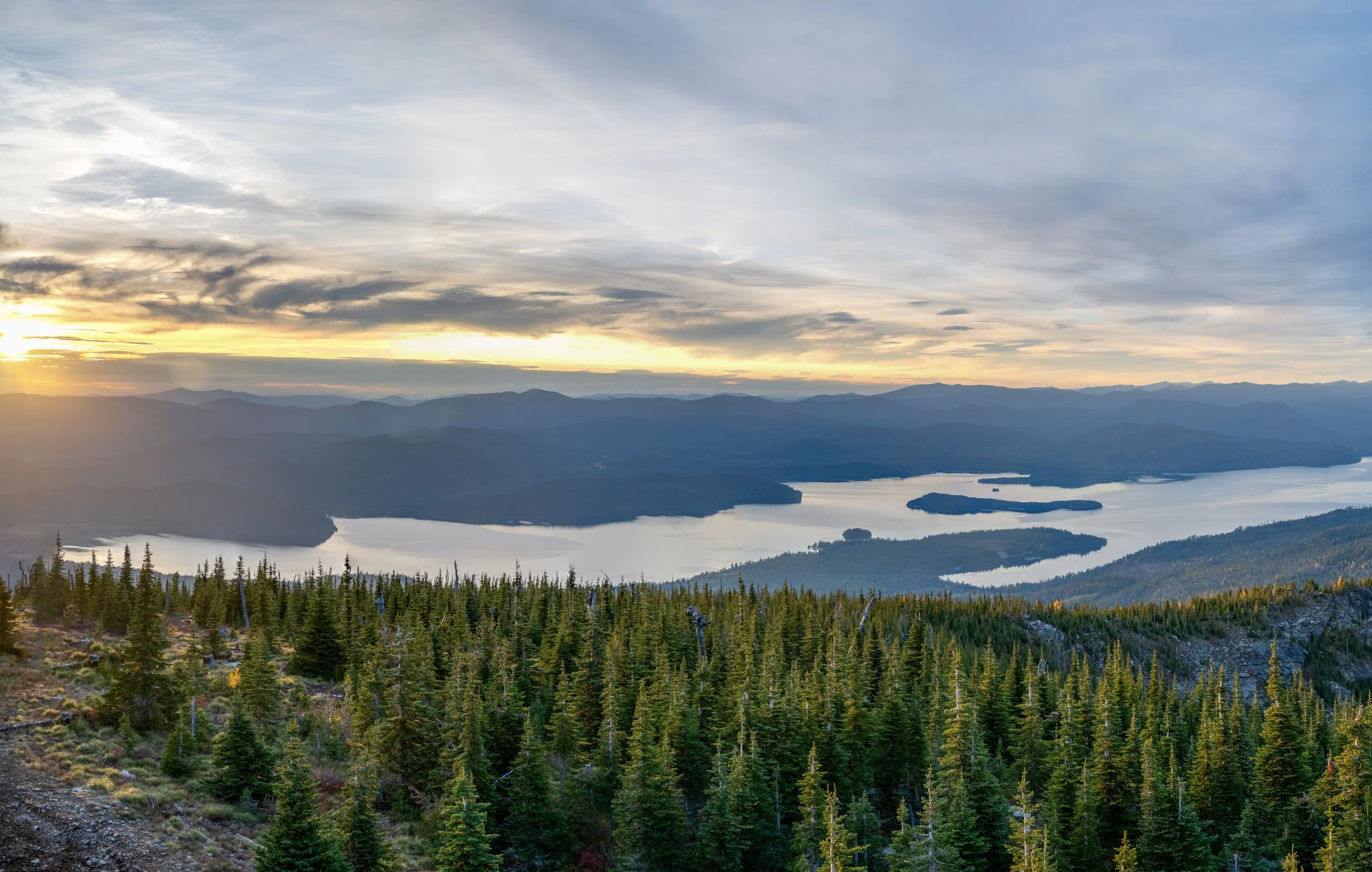 Sunset over a mountain landscape with a large body of water, surrounded by forested hills in the foreground.