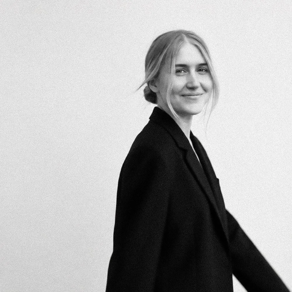 Black and white portrait of a woman with blonde hair tied back, smiling, wearing a dark blazer, against a plain light background.