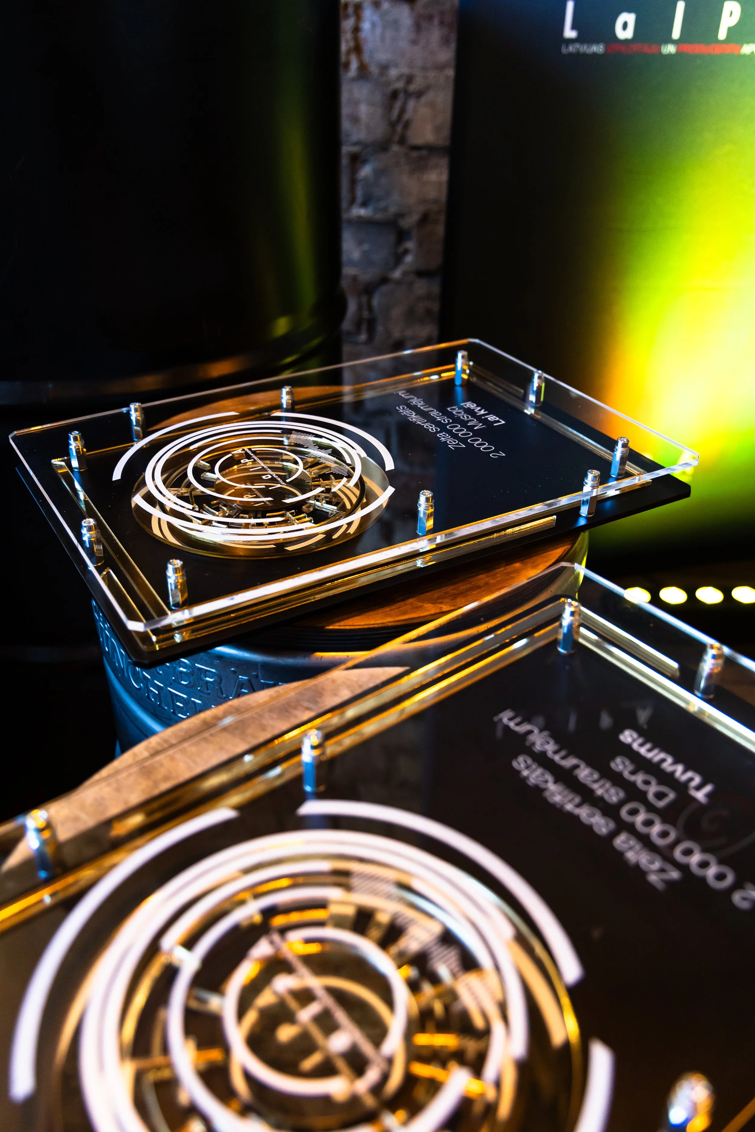 Close-up of a transparent acrylic award or plaque with engraved text, sitting on top of a tin can with a spiral pattern, with dark background and blurry vent or appliance with control panel.
