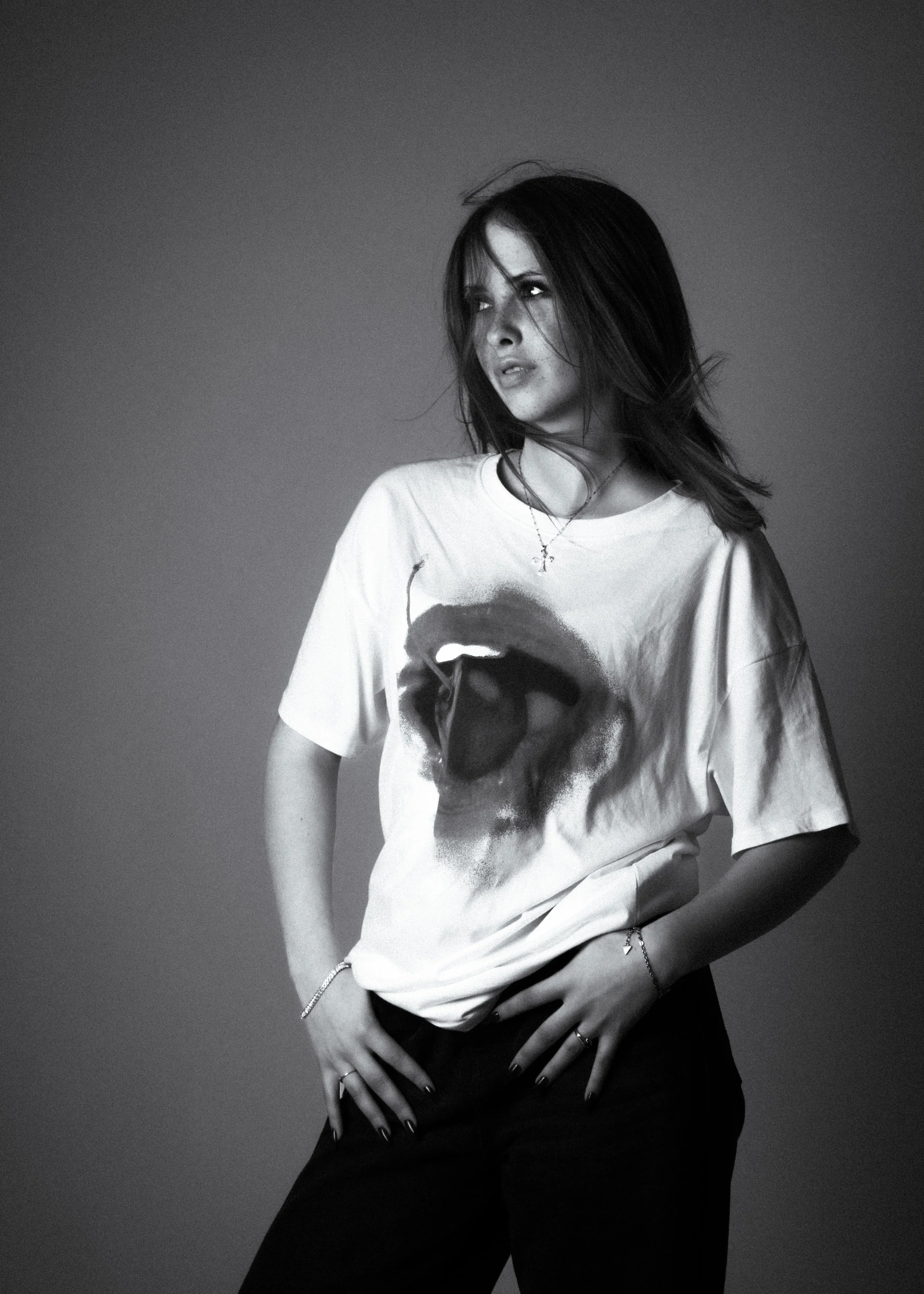 Black and white photo of a young woman with dark hair, wearing a white t-shirt with a face printed on it, black pants, and jewelry, standing against a plain background.