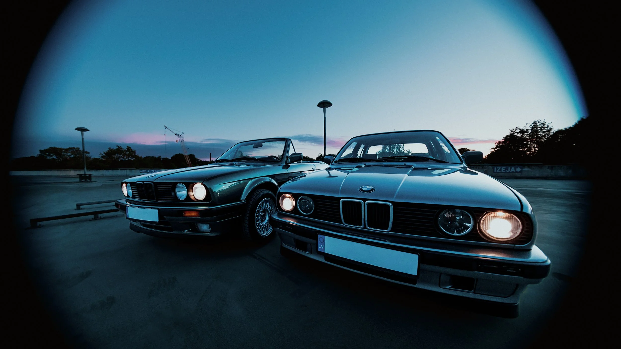 Two classic BMW cars parked on a rooftop parking lot during dusk, with a construction crane and trees in the background.