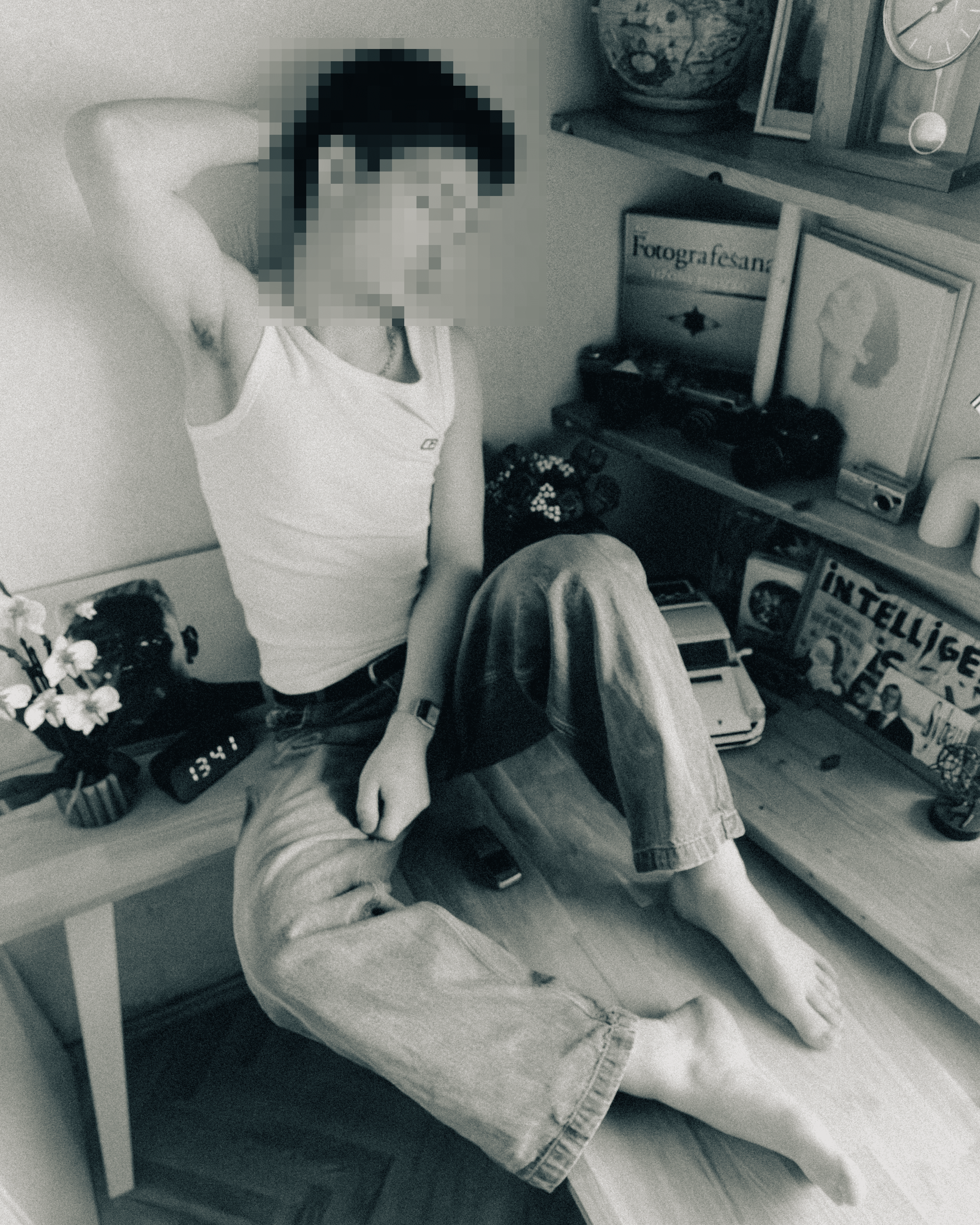 A person with short dark hair, wearing a tank top, relaxing with arms behind head, sitting on a table with books, magazines, framed artwork, and a clock on the shelf nearby.
