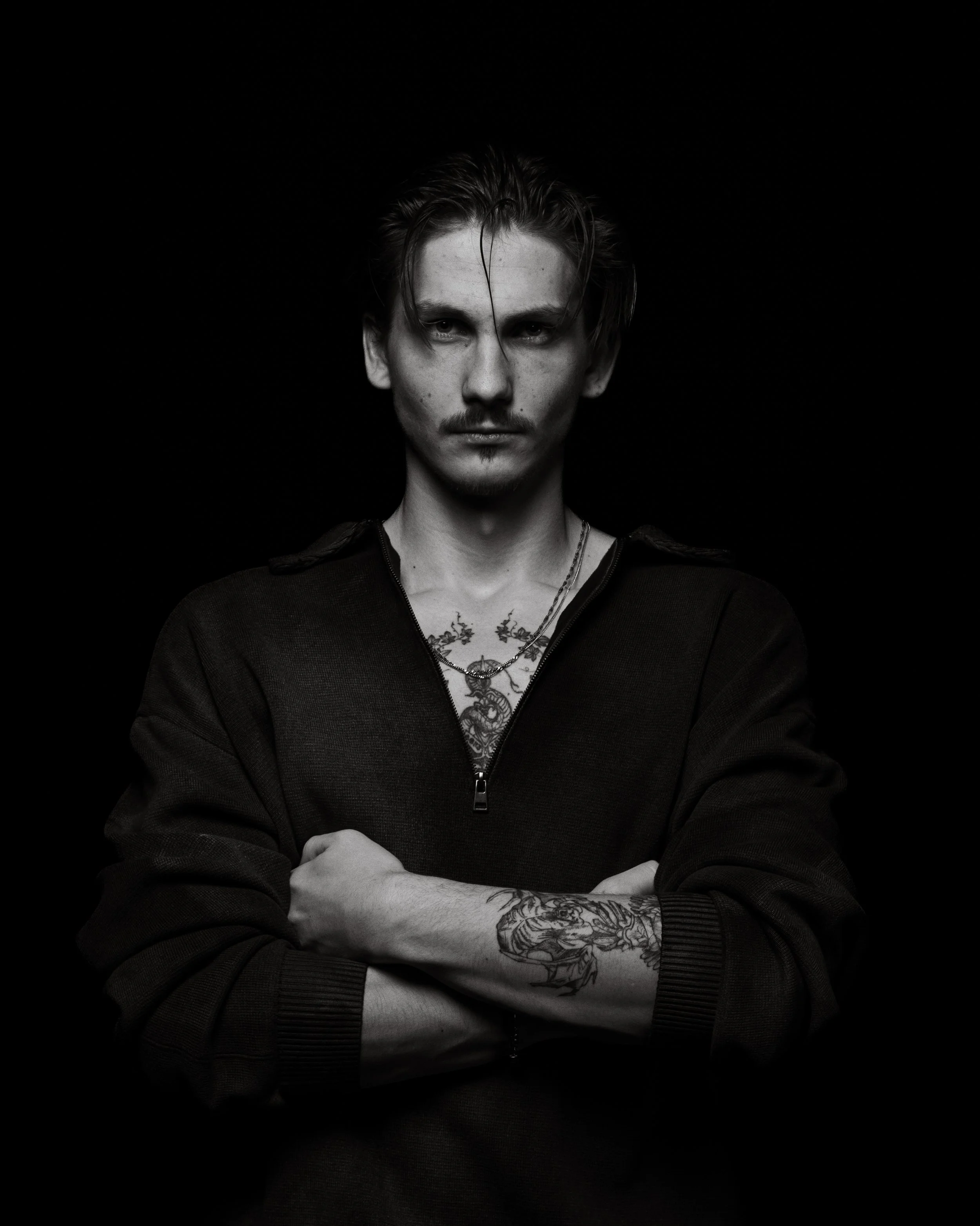 A black and white portrait of a young man with short, slicked-back hair, looking directly at the camera with a serious expression. He has facial tattoos and tattoos on his arms and chest, and is wearing a dark zipped top and layered necklaces. His ar
